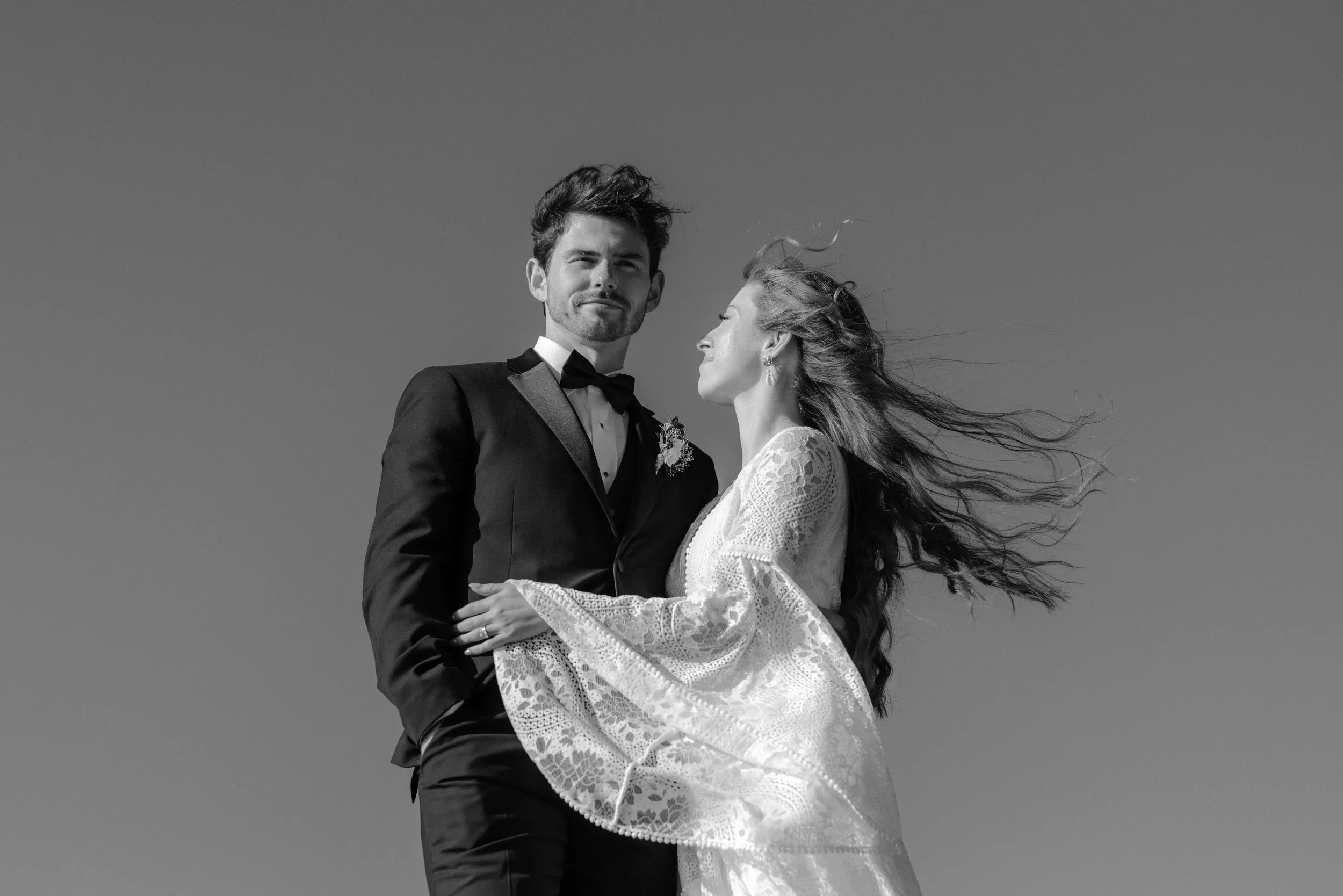 A black and white photograph of a bride and groom standing outdoors against a clear sky. The groom is wearing a tuxedo with a bow tie, and the bride is in a lace wedding dress. The bride's hair is flowing in the wind, and she is looking at the groom, who is looking confidently at the camera.