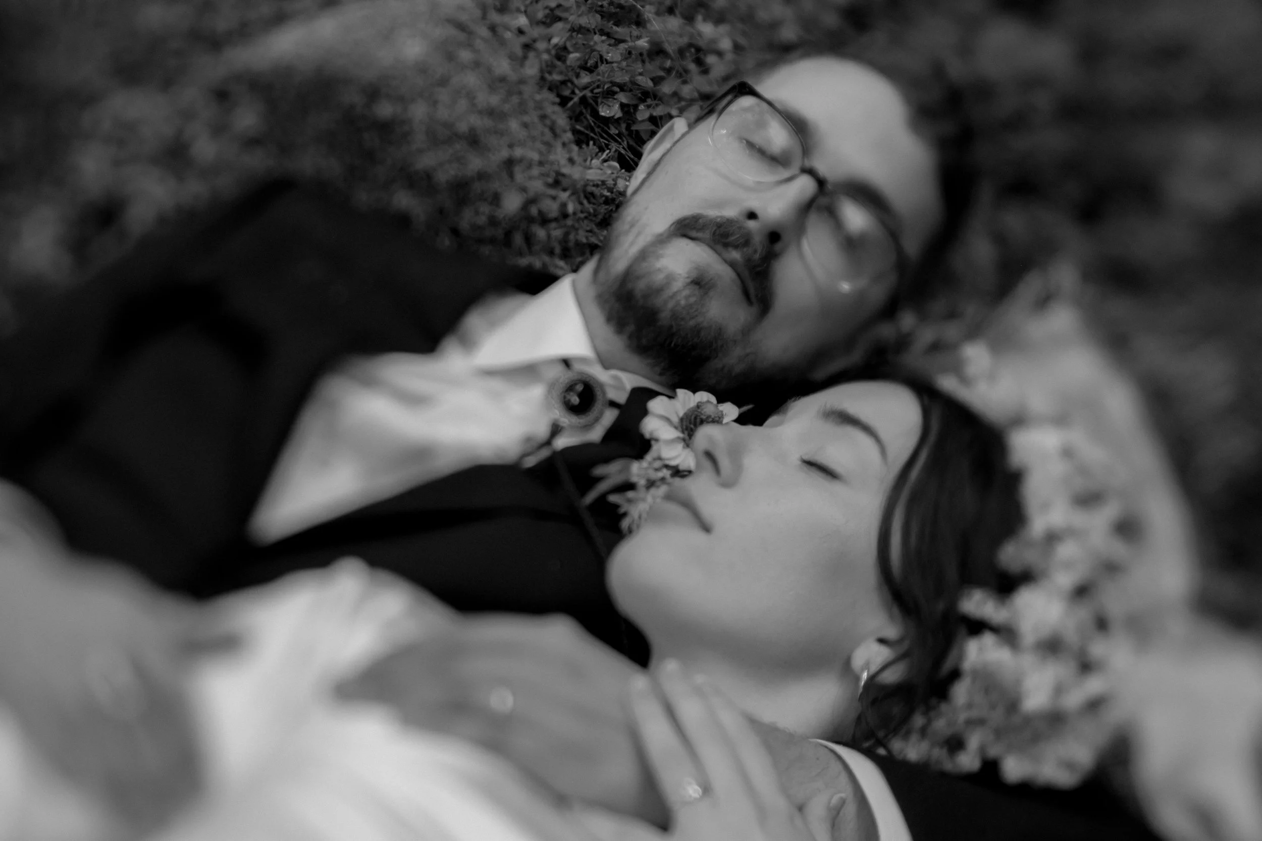 Black and white photo of a man and woman lying on the ground, close to each other, eyes closed, with their faces gently touching. The woman has short dark hair, and the man has glasses and facial hair. The scene appears peaceful and intimate.