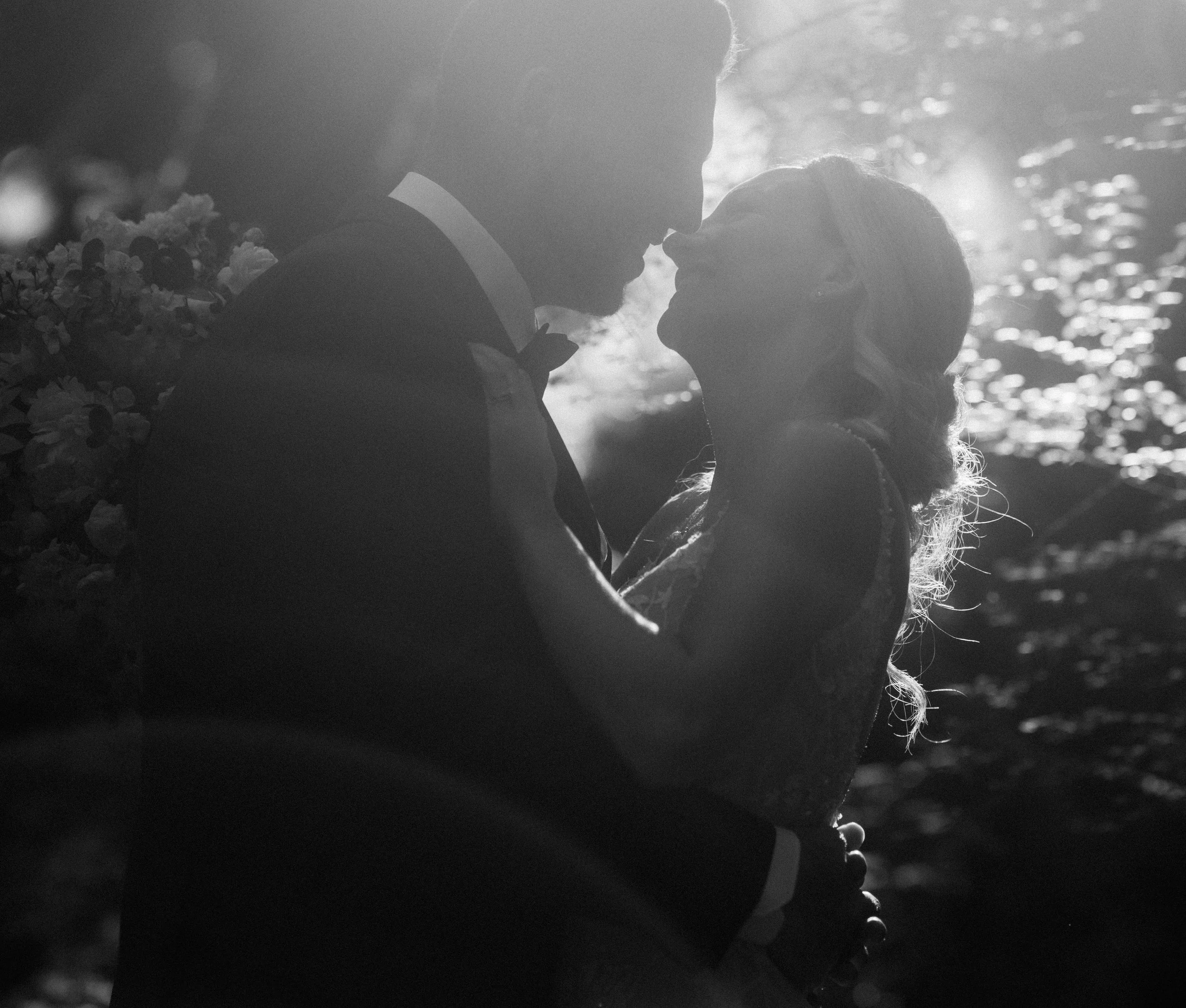 A silhouette of a newlywed couple, with the groom holding the bride close, standing near a body of water with sunlight reflecting off it.