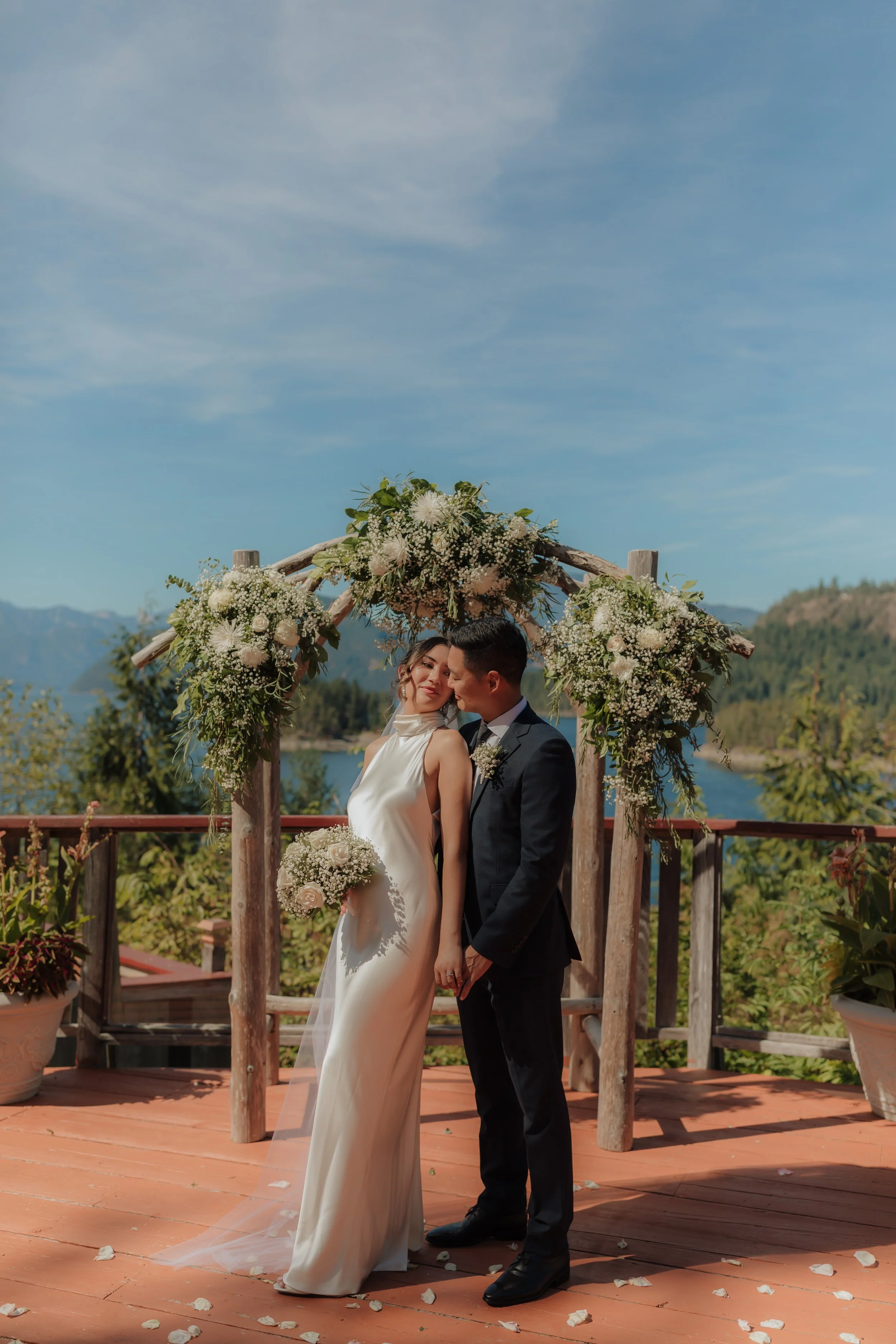 A newlywed couple holding hands and smiling at each other under a floral wedding arch on an outdoor wooden deck with a scenic mountain and water background.
