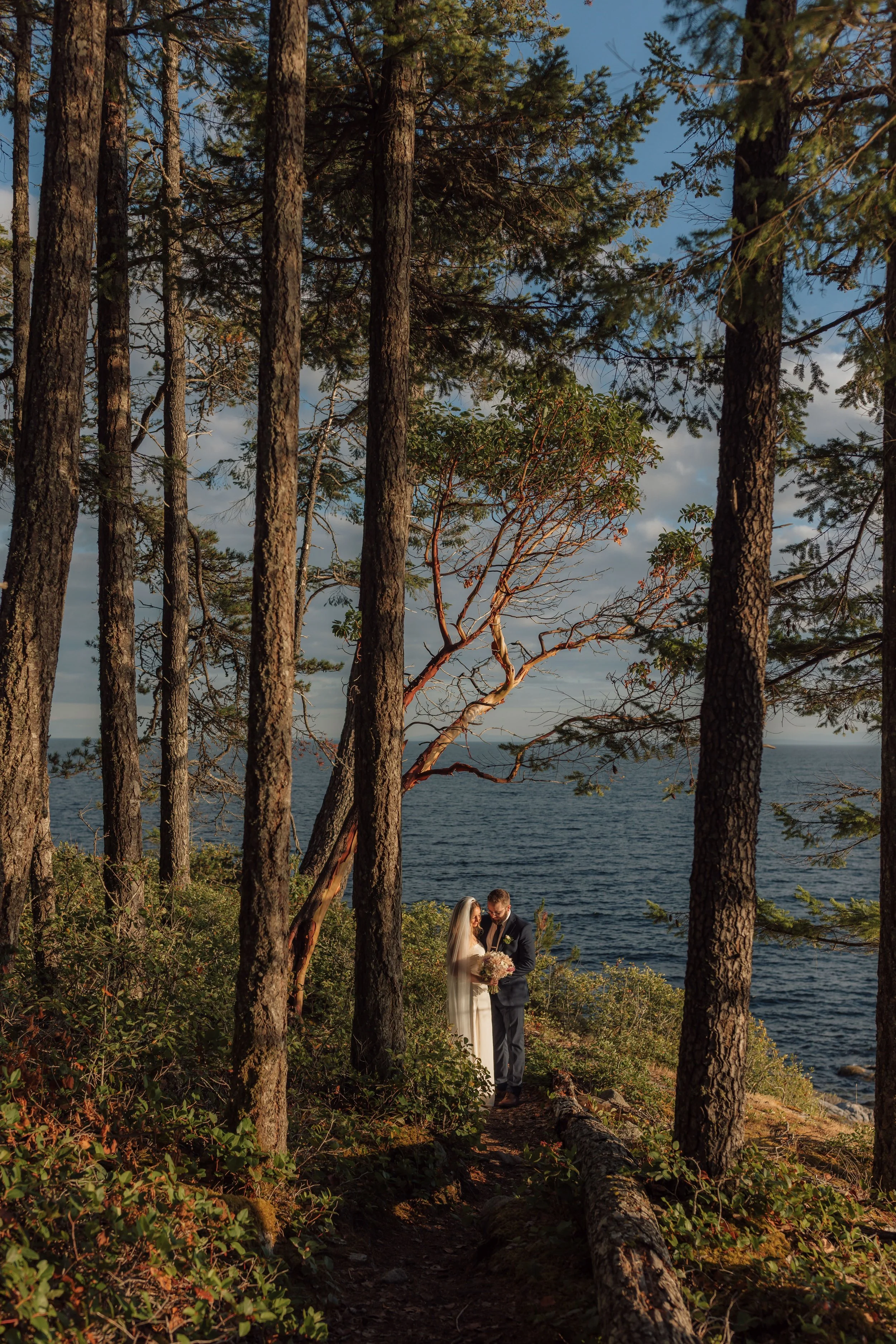A bride and groom standing on a trail in a forest near the water, framed by tall trees, during sunset.