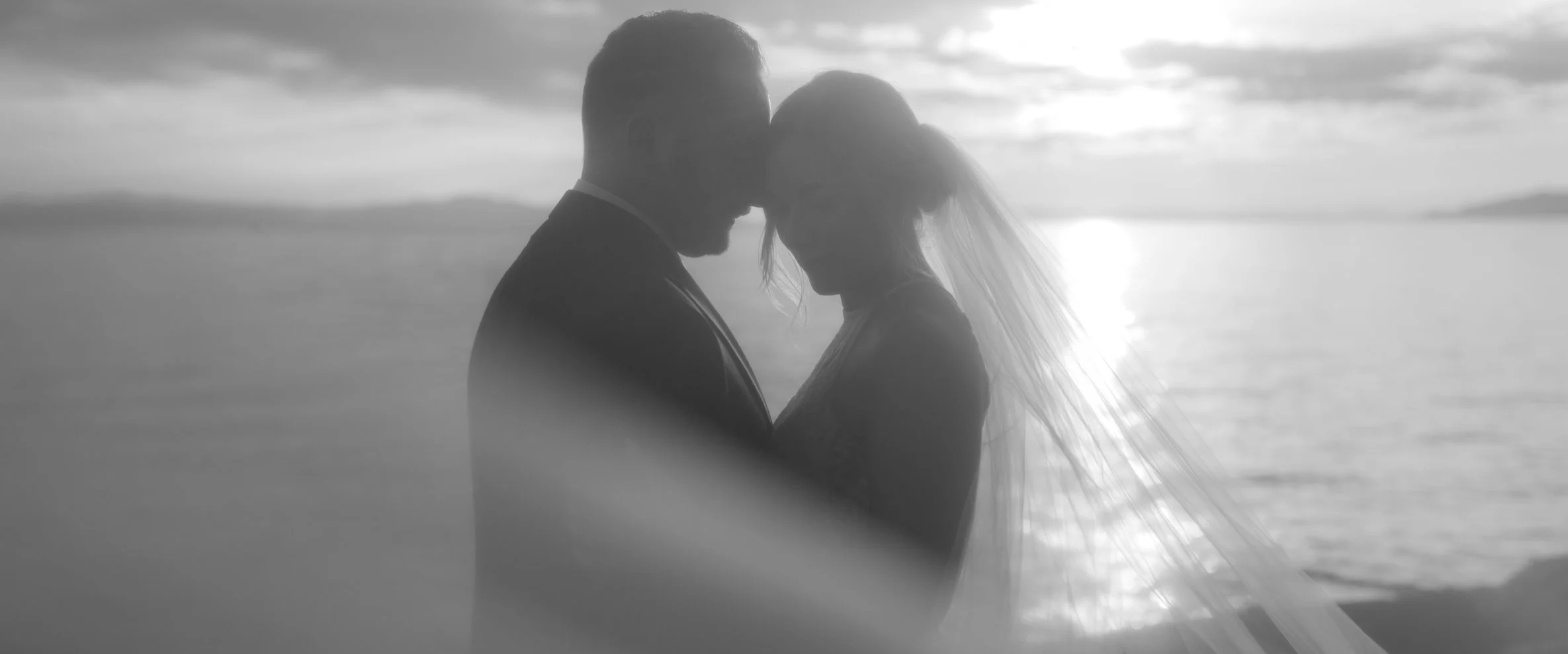 A black and white photo of a bride and groom embracing by the water at sunset or sunrise, with their foreheads touching.