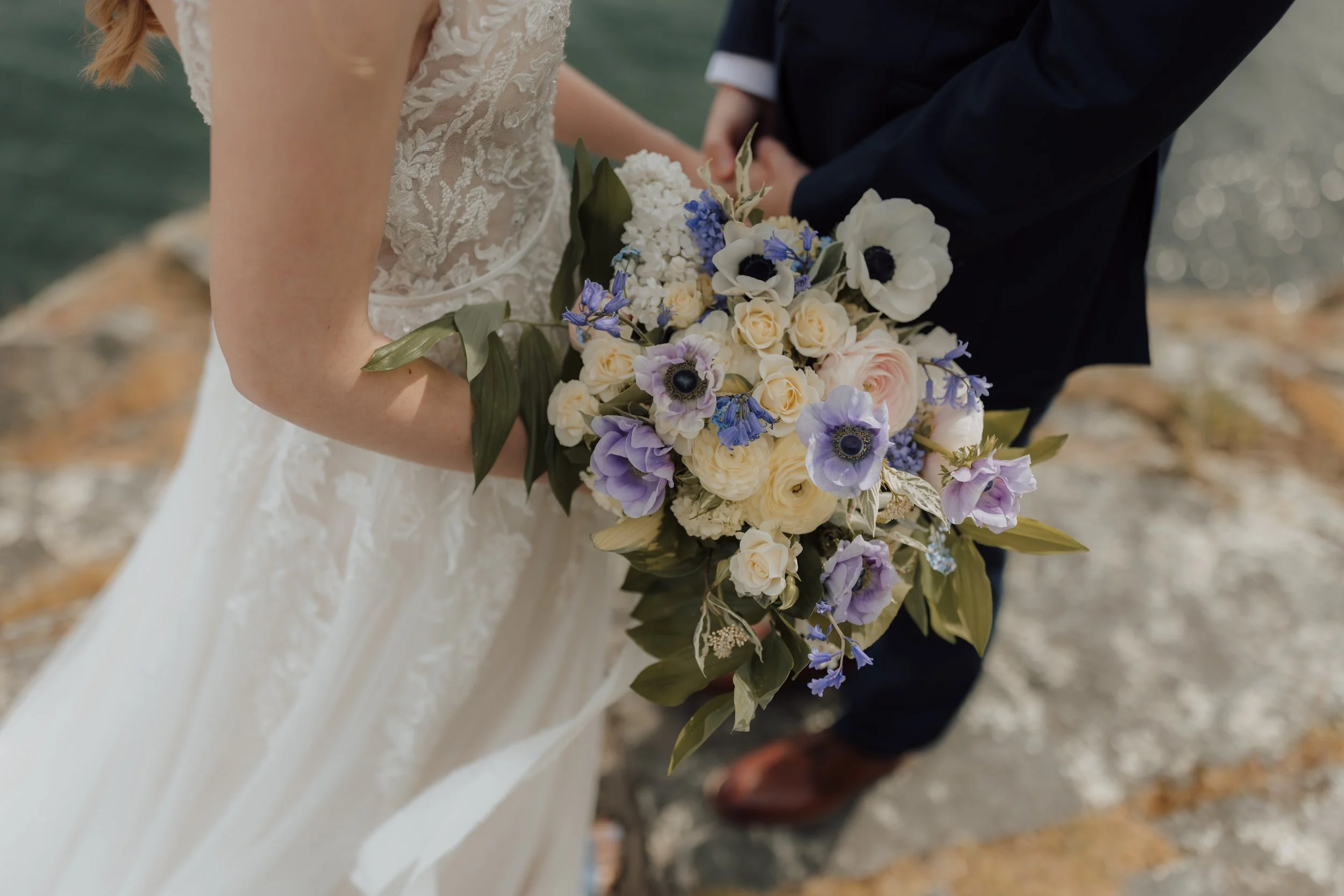 A bride and groom holding a bouquet of white, lavender, and blue flowers during their wedding at the beach at their elopement in Gibsons.