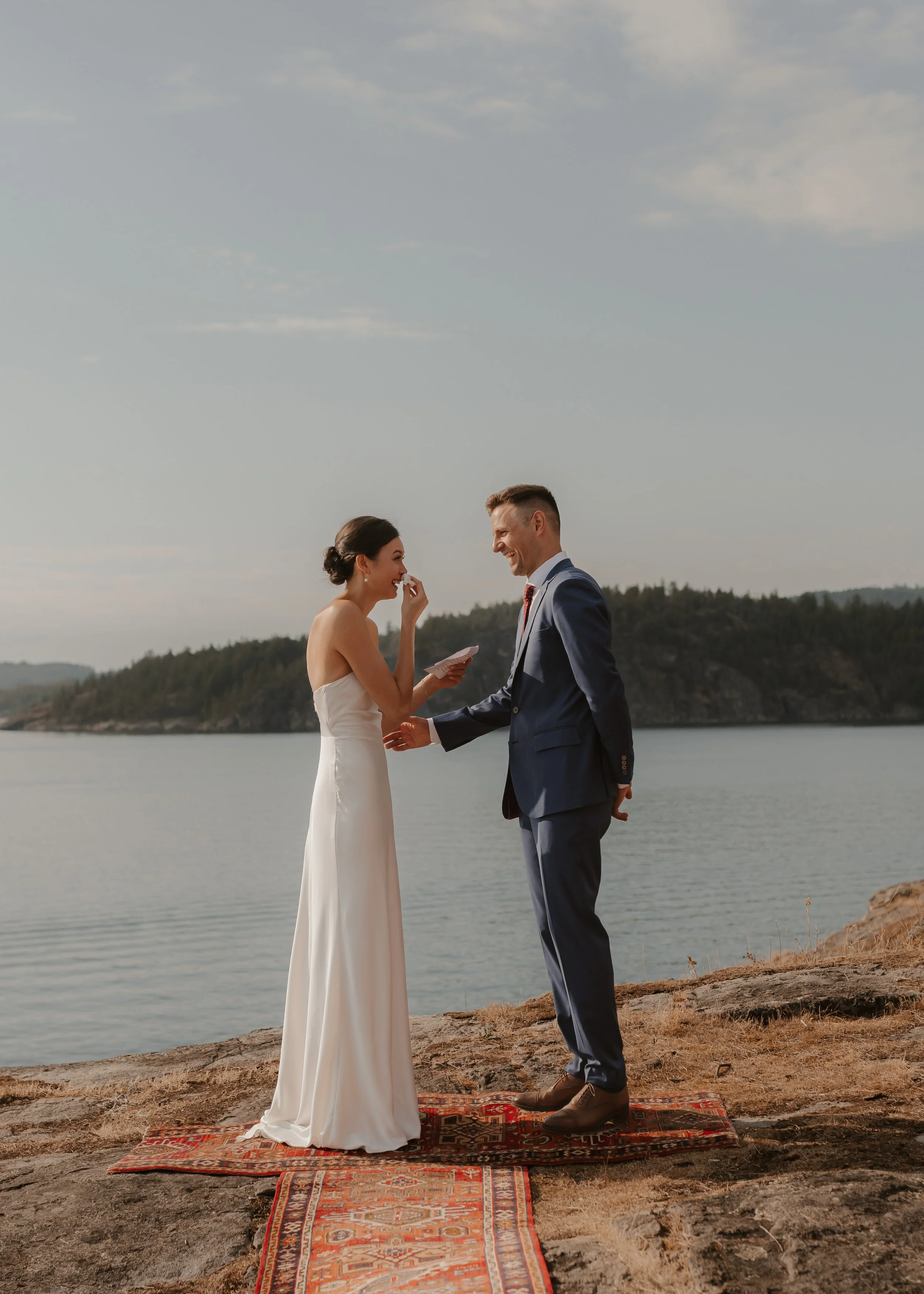 A couple stands on a red patterned rug at the edge of a body of water, exchanging vows with a scenic landscape in the background.