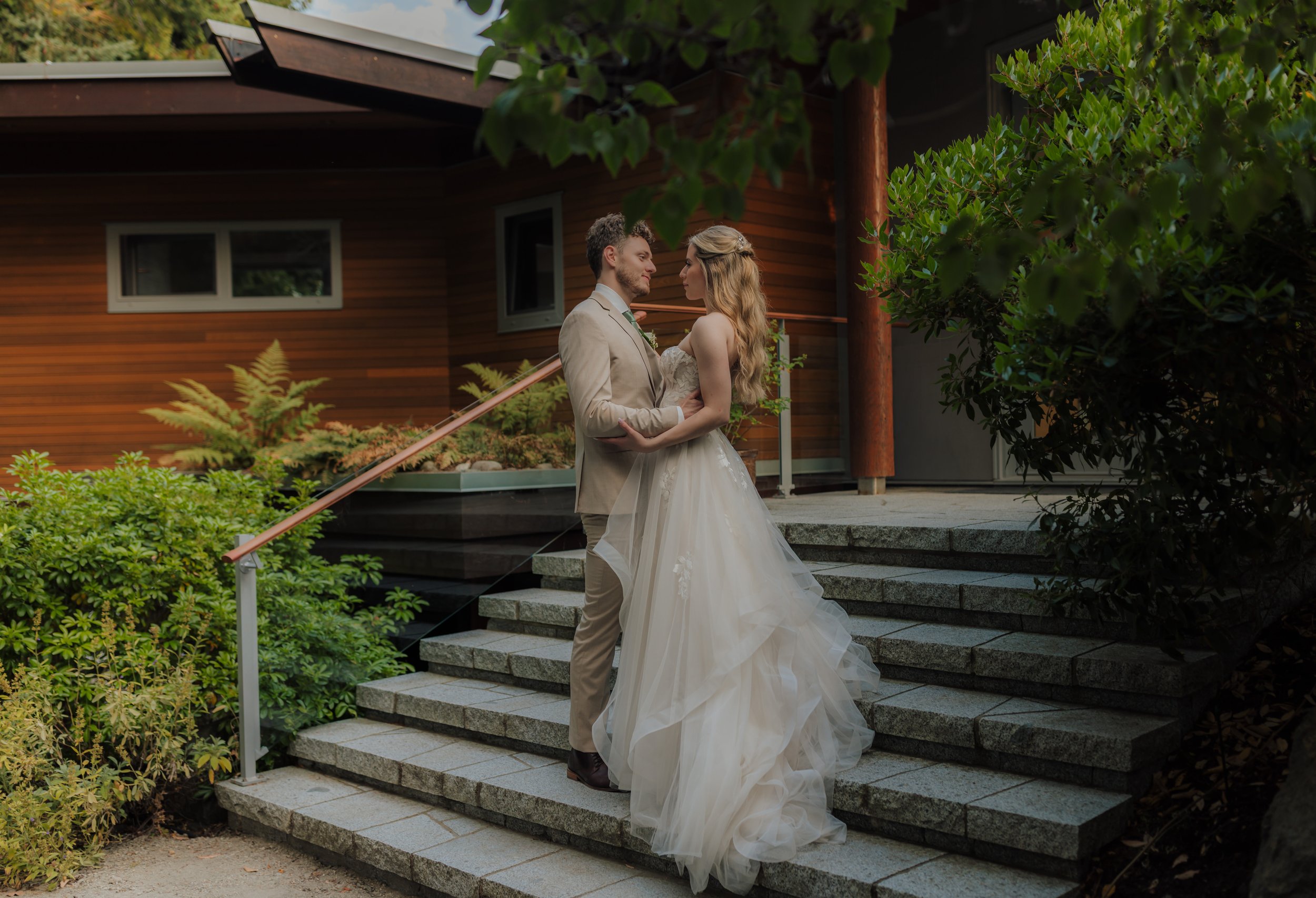 A bride and groom stand on outdoor stone stairs, gazing into each other's eyes, with a wooden building and greenery in the background at the Pointhouse on the Sunshine Coast.