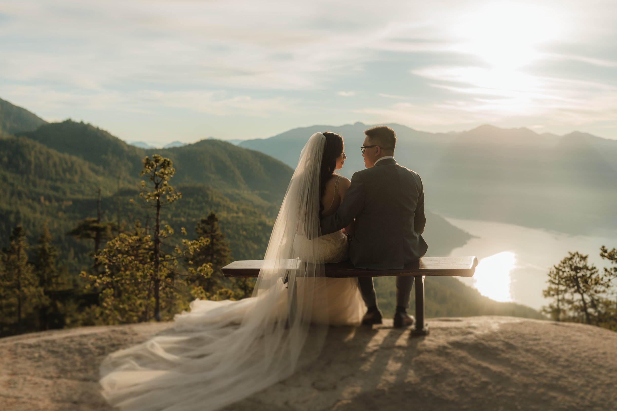 A bride and groom sitting on a bench overlooking a scenic mountain landscape at sunset at the Sea to Sky Gondola.