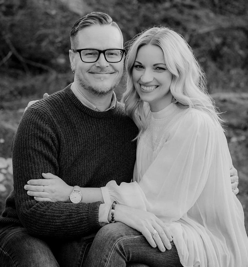 A black and white photo of a smiling couple sitting close together outdoors, embracing each other. The man is wearing glasses, a dark sweater, and has short, styled hair. The woman has long, wavy hair, and is wearing a light-colored blouse or dress. They are smiling happily at the camera.