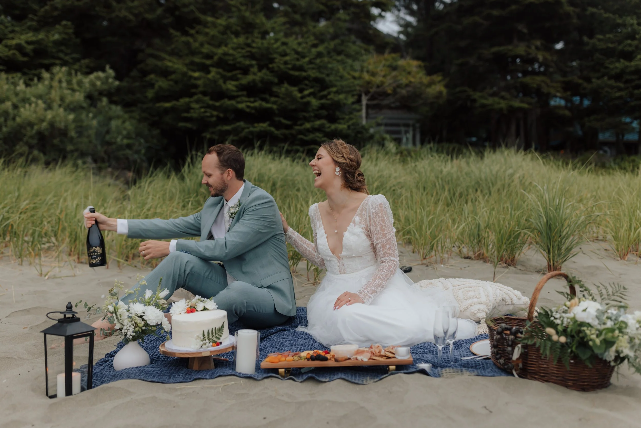 Bride and groom celebrating on a beach, sitting on a blanket with food and flowers, bride wearing a white lace dress, groom in a gray suit, beach setting with grasses, trees, and outdoor decor on Chesterman Beach in Tofino, BC.