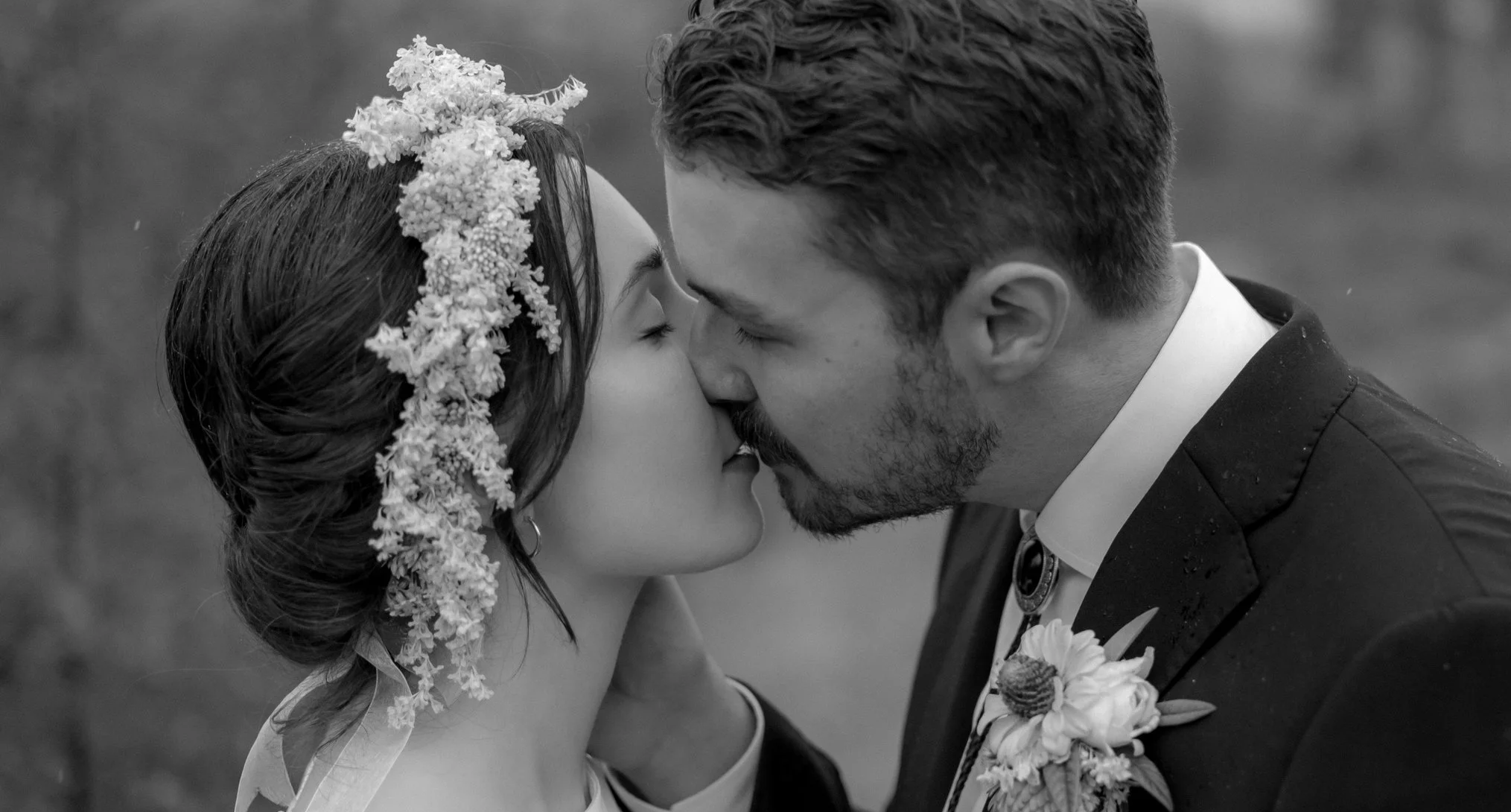A black-and-white photo of a bride and groom kissing outdoors, with the bride wearing a flower crown and the groom wearing a boutonniere.