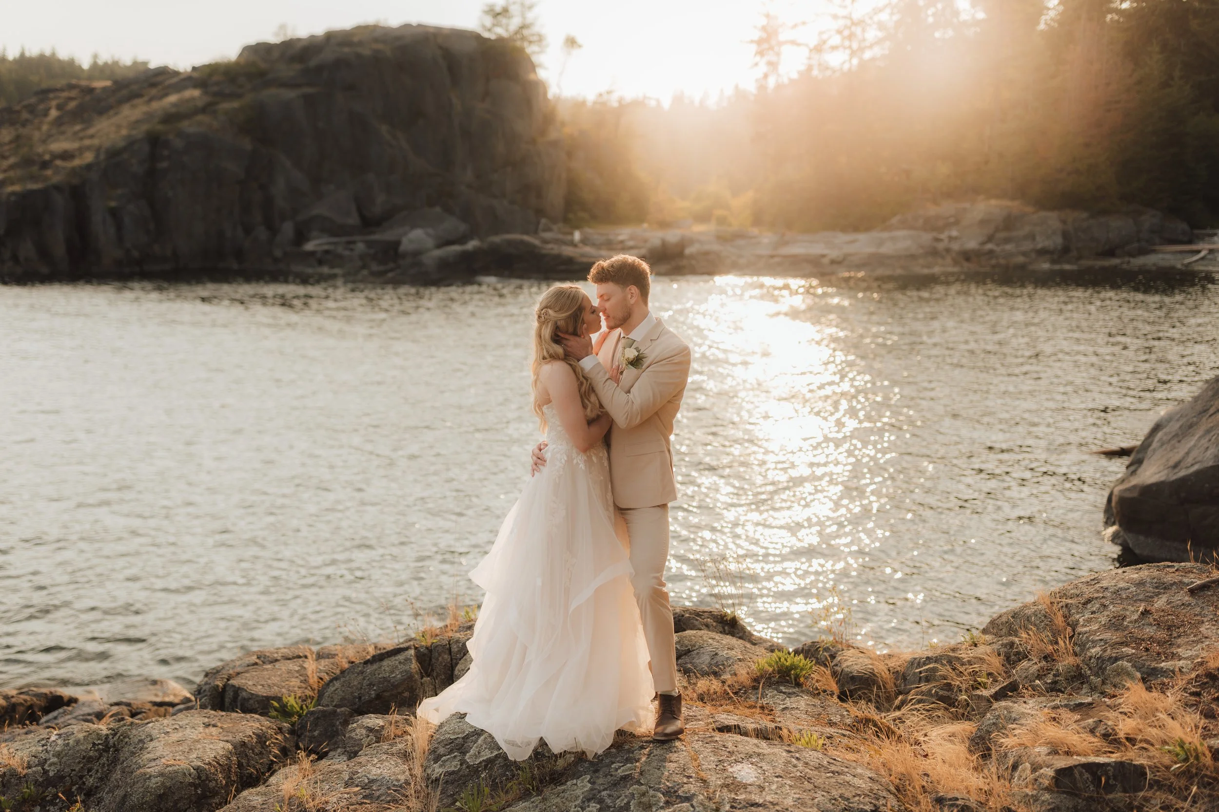 A bride and groom standing on rocks by the ocean during sunset, embracing and gazing at each other at the Pointhouse on the Sunshine Coast.