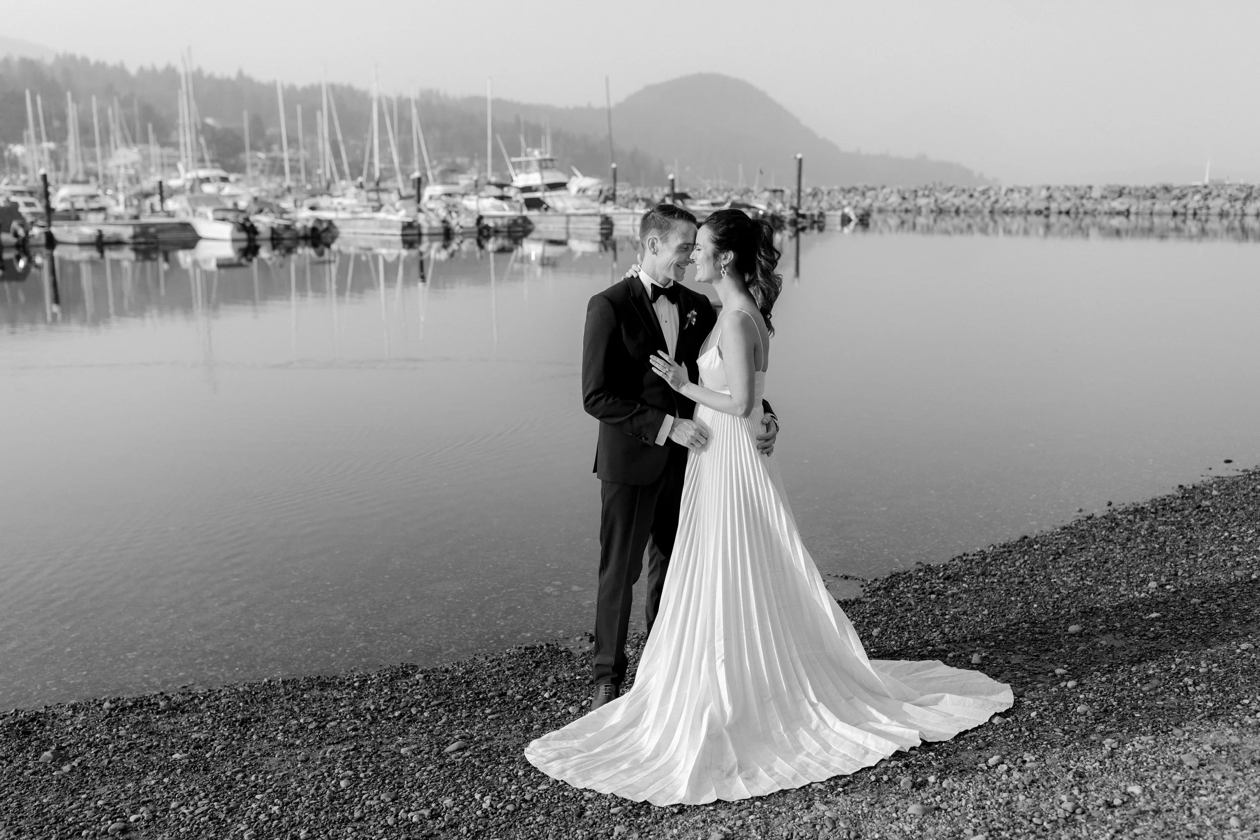 A Black and white photo of a wedding couple standing on a rocky shoreline by a marina, with boats and a mountain in the background. The bride is wearing a flowing pleated wedding gown, and the groom is in a tuxedo. In Gibsons on the Sunshine Coast.
