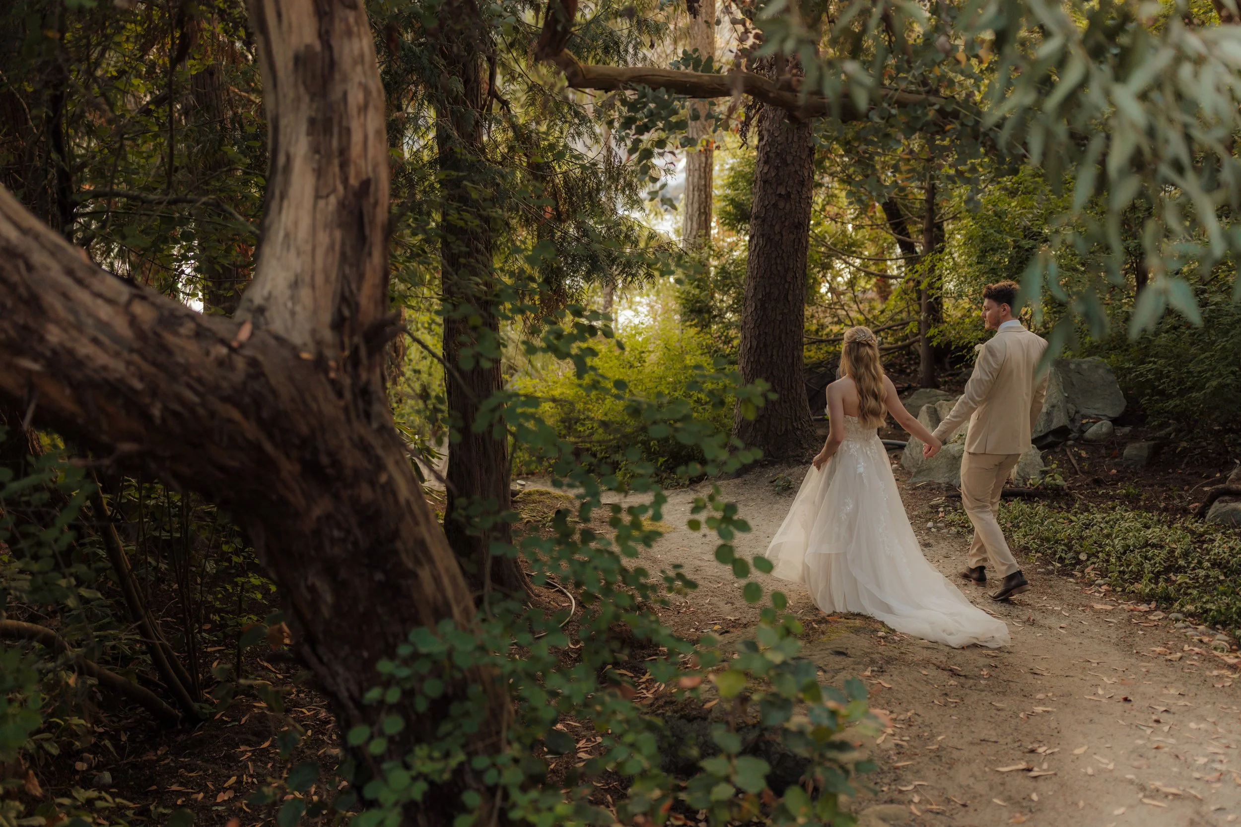 A bride and groom walking hand in hand along a wooded trail in a forest setting, surrounded by trees and greenery at the Pointhouse on the Sunshine Coast.
