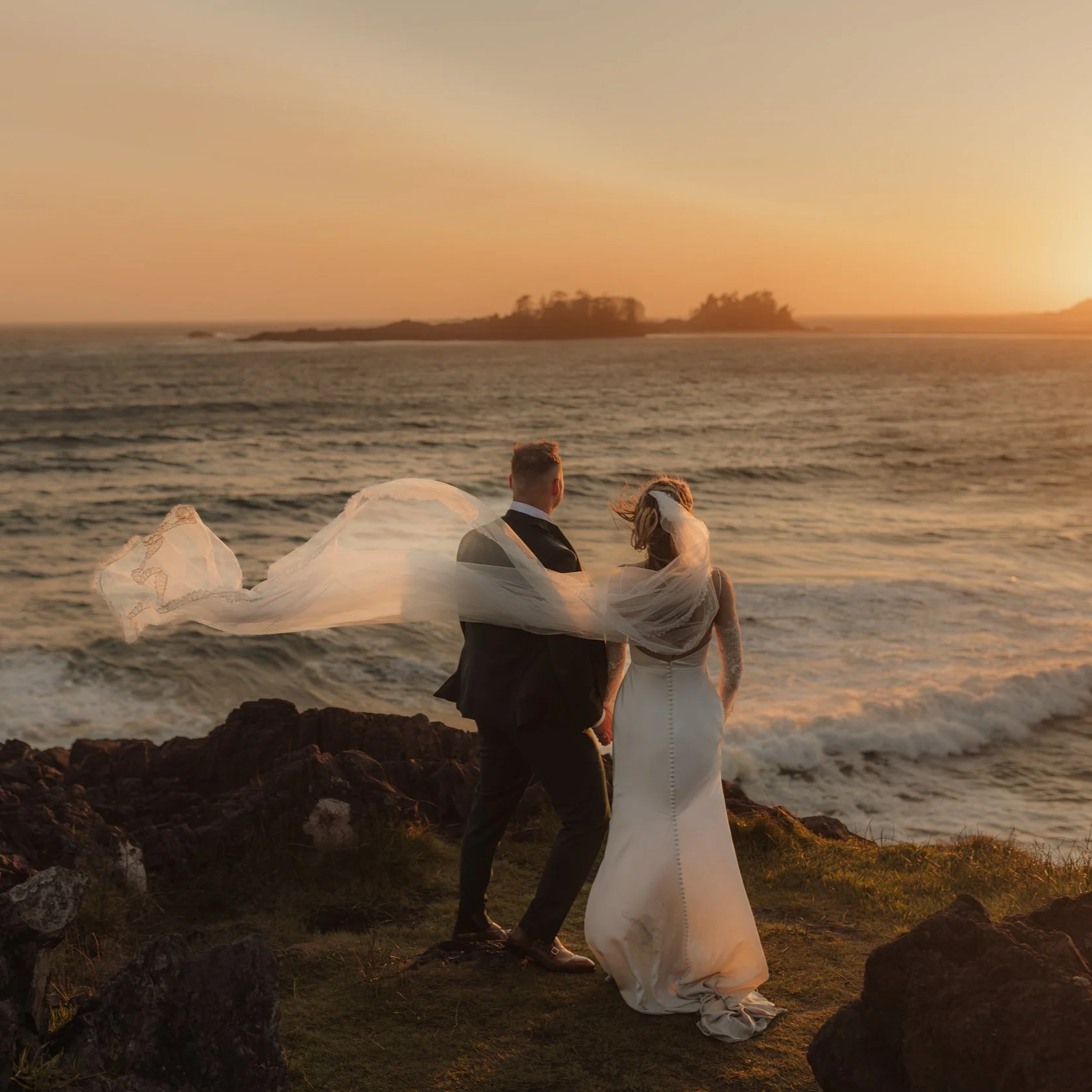 A bride and groom holding hands on a rocky shoreline during sunset, with the bride's veil flowing in the wind and the ocean in the background at Pettinger Point in Tofino, BC.
