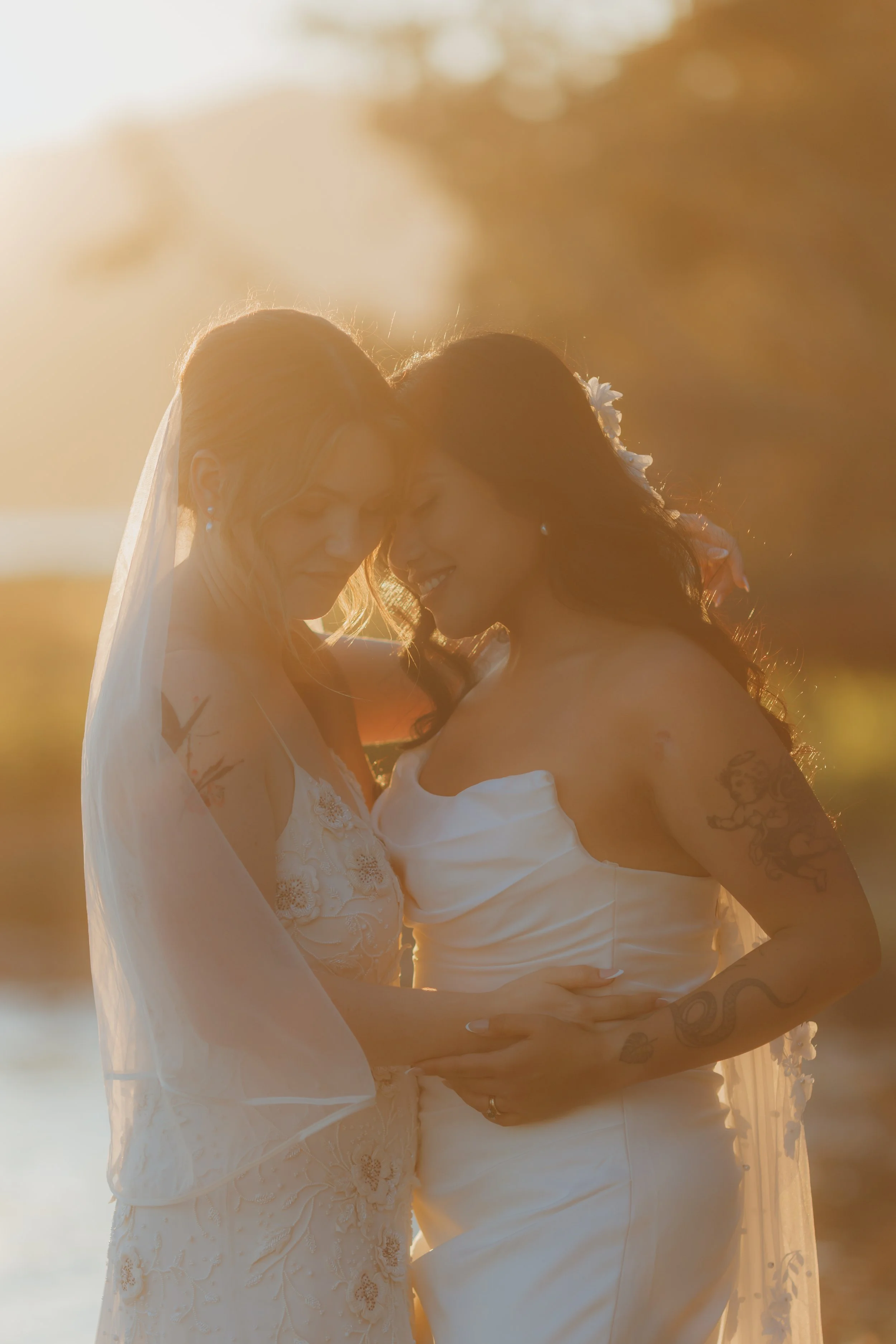 Two women in wedding dresses share an intimate moment at sunset.