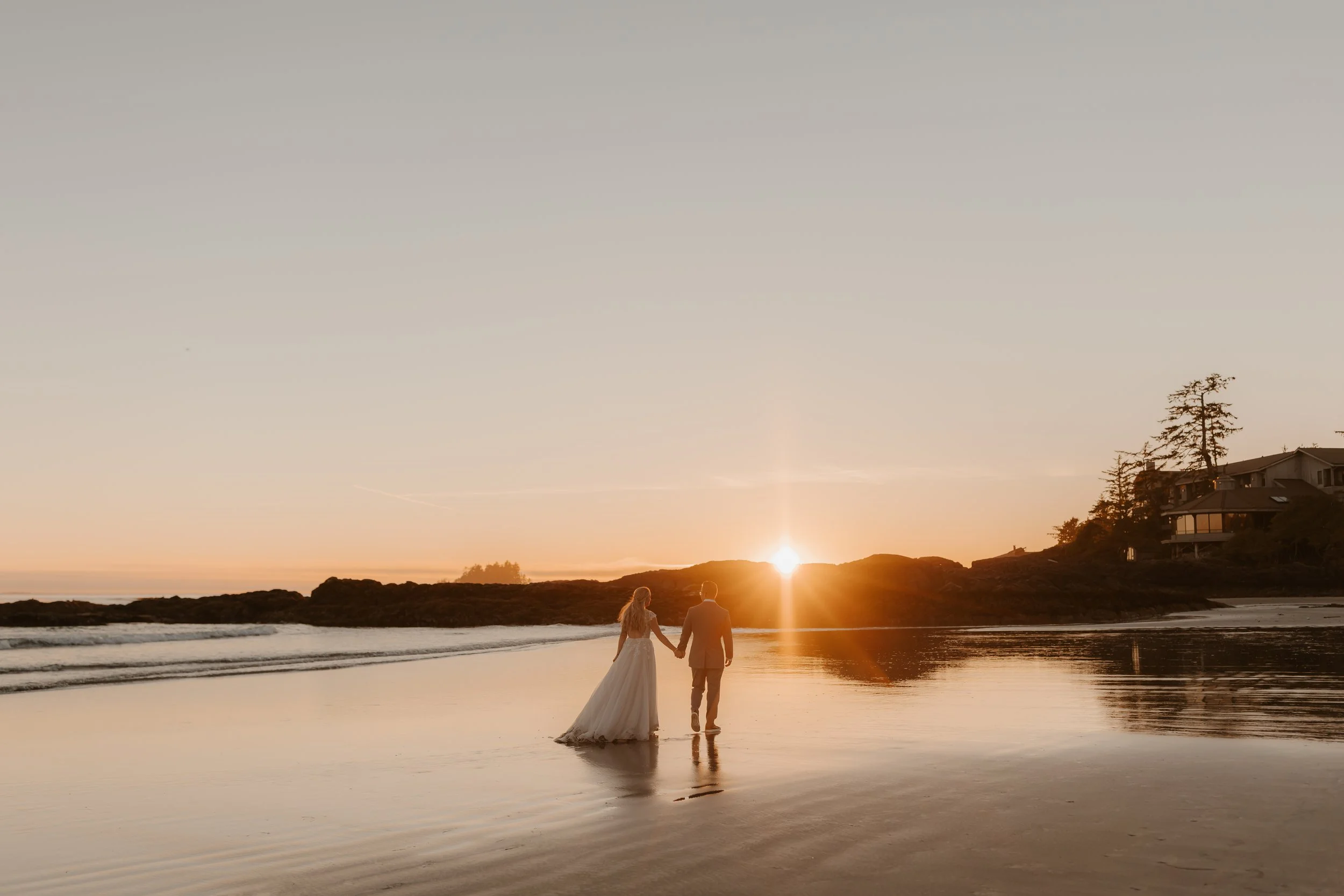 A couple in wedding attire walking hand in hand along the shoreline at sunset, with the bride in a white gown and the groom in a suit, reflecting in the wet sand on the beach.