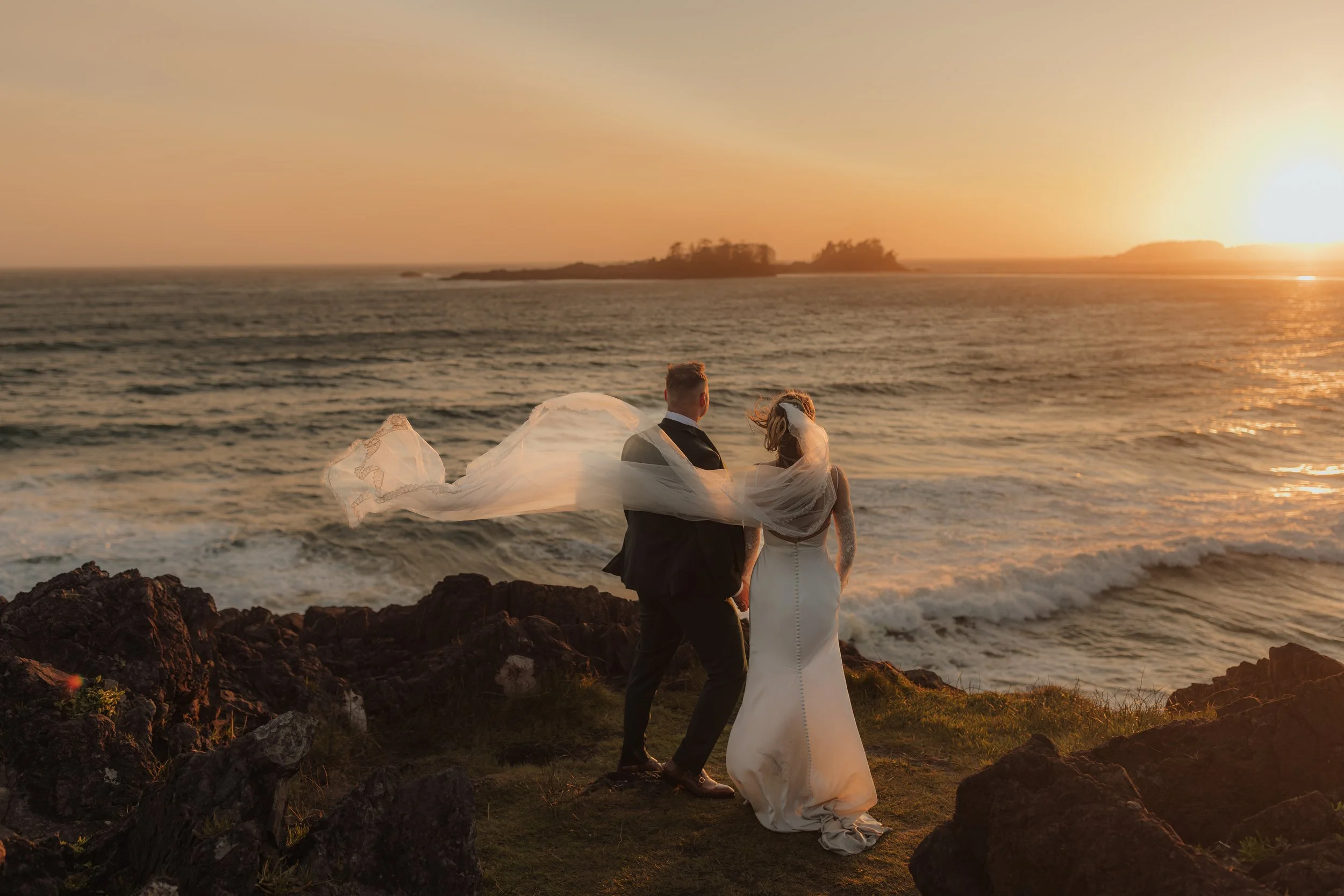 A bride and groom holding hands on a rocky coastline at sunset, with the bride’s veil flowing in the wind and the ocean waves in the background at Pettinger Point