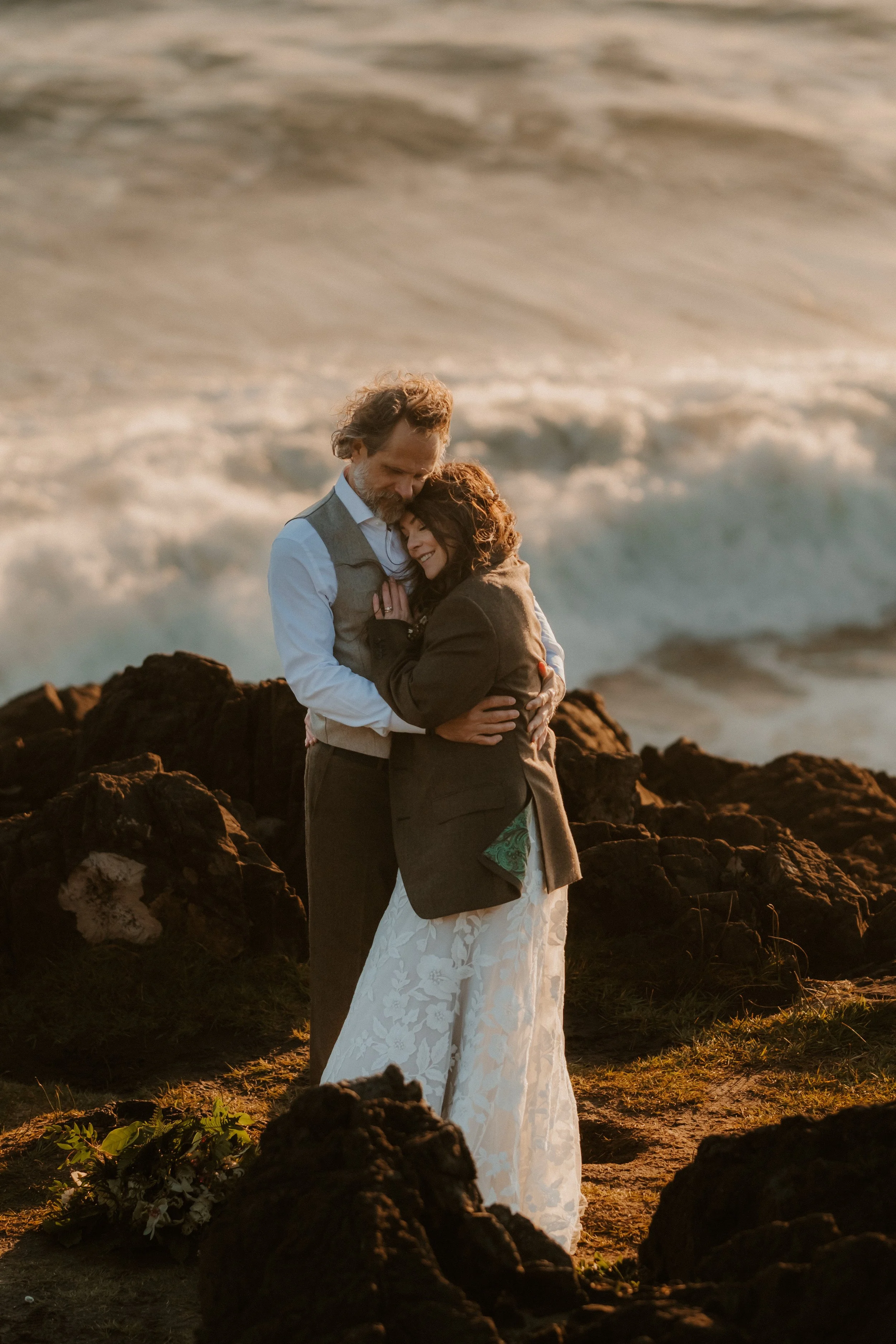 A couple embracing on a rocky beach at sunset, with ocean waves in the background at Pettinger Point in Tofino, BC
