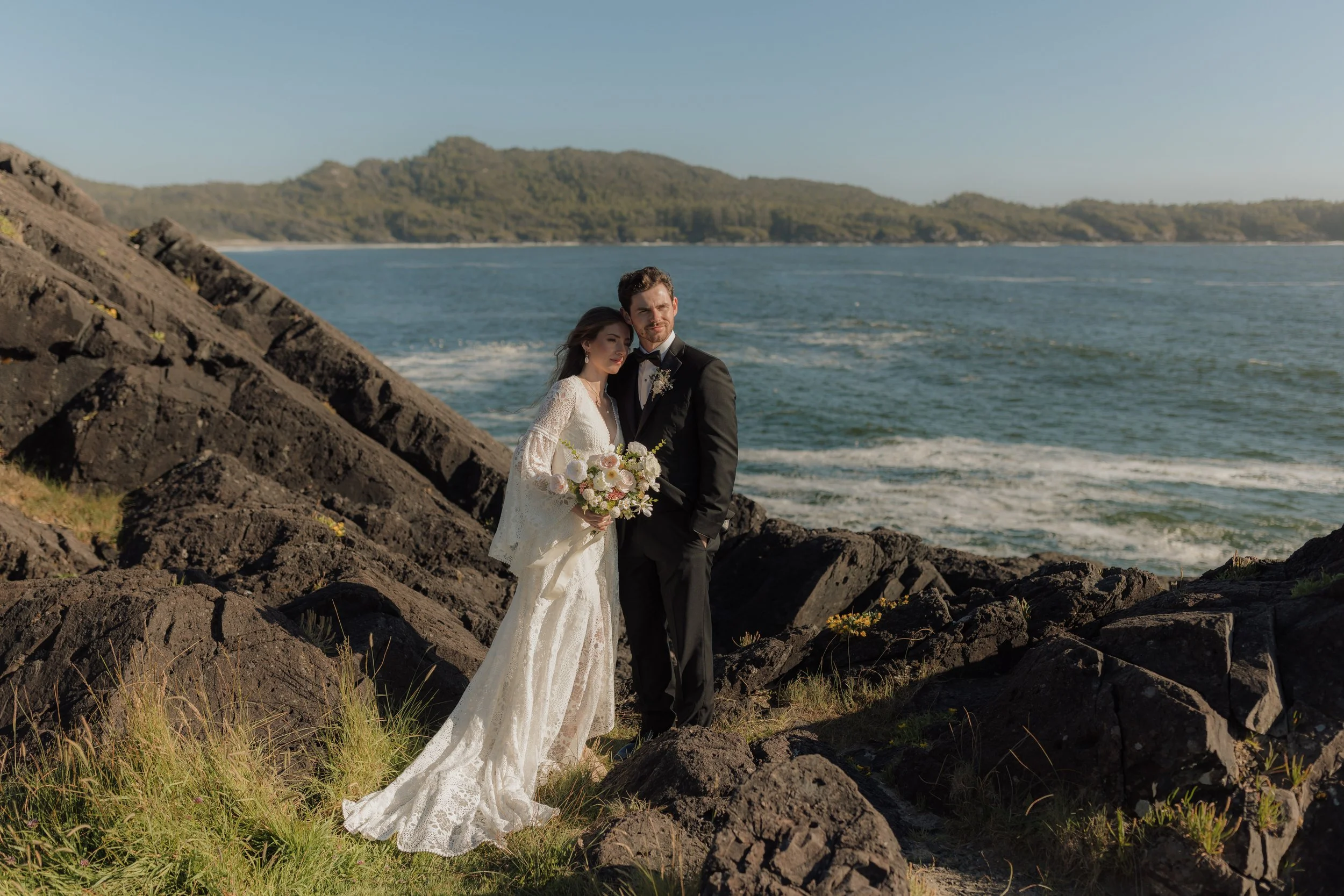 A bride and groom standing on rocks near the ocean with green hills in the background, during their wedding day at Pettinger Point in Tofino.