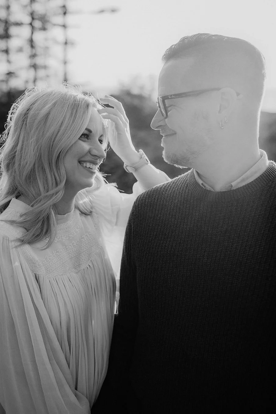 A black and white photo of a smiling woman with wavy blonde hair and a man with glasses and a beard looking at each other outdoors, with trees in the background.