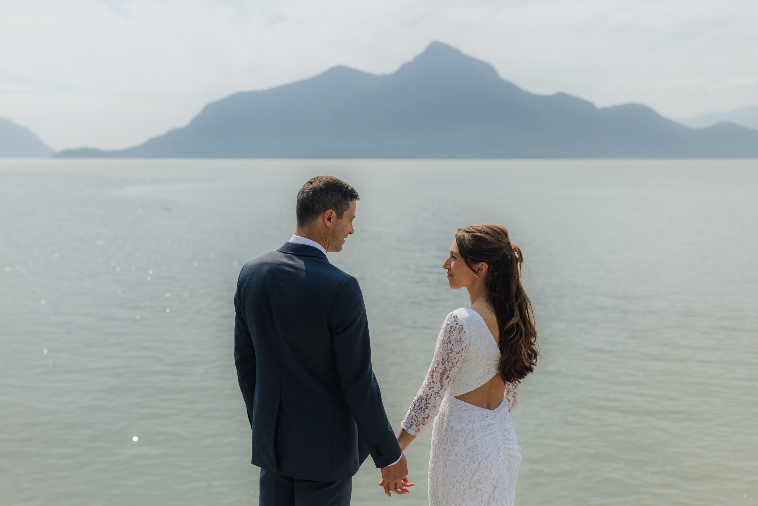 A bride and groom standing by the water holding hands with mountains in the background, during a wedding or romantic photo session at Porteau Cove in Squamish, BC.