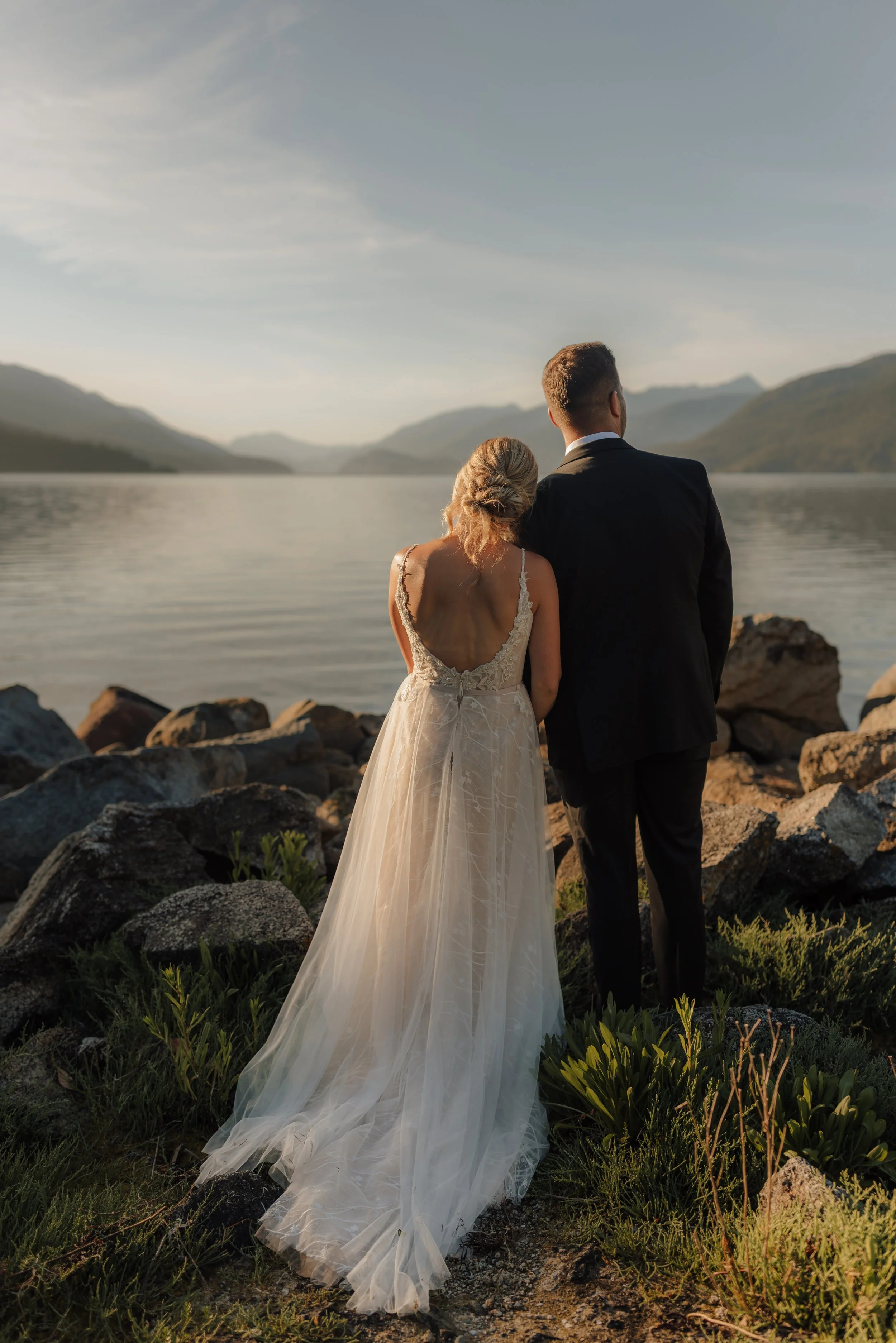 Couple dressed in wedding attire standing by the ocean with mountains in the background at sunset on the Sunshine Coast.