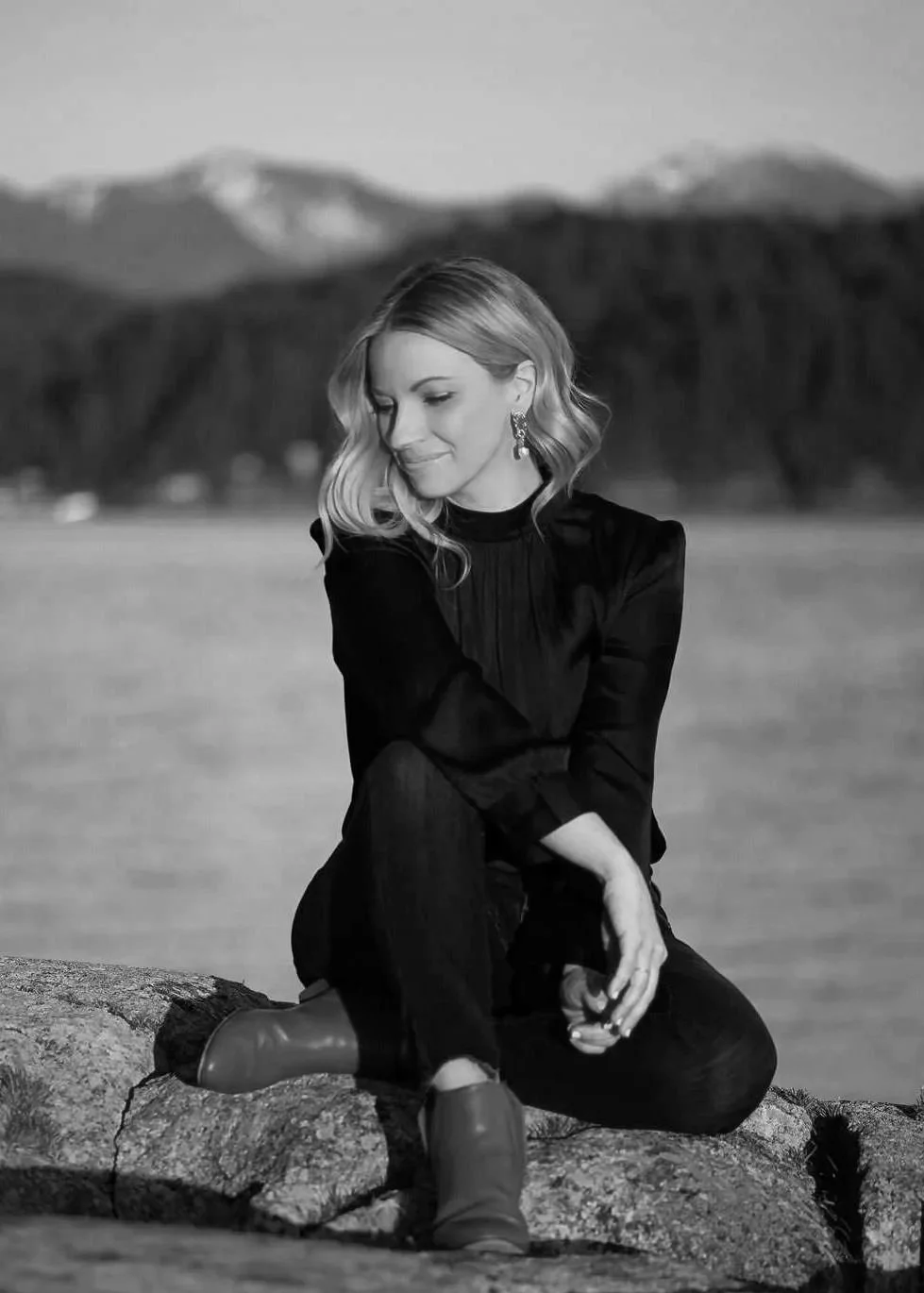 A woman sitting on a rock by the water, with mountains in the background, captured in black and white. She is looking down with a gentle smile, wearing earrings and a dark blouse.