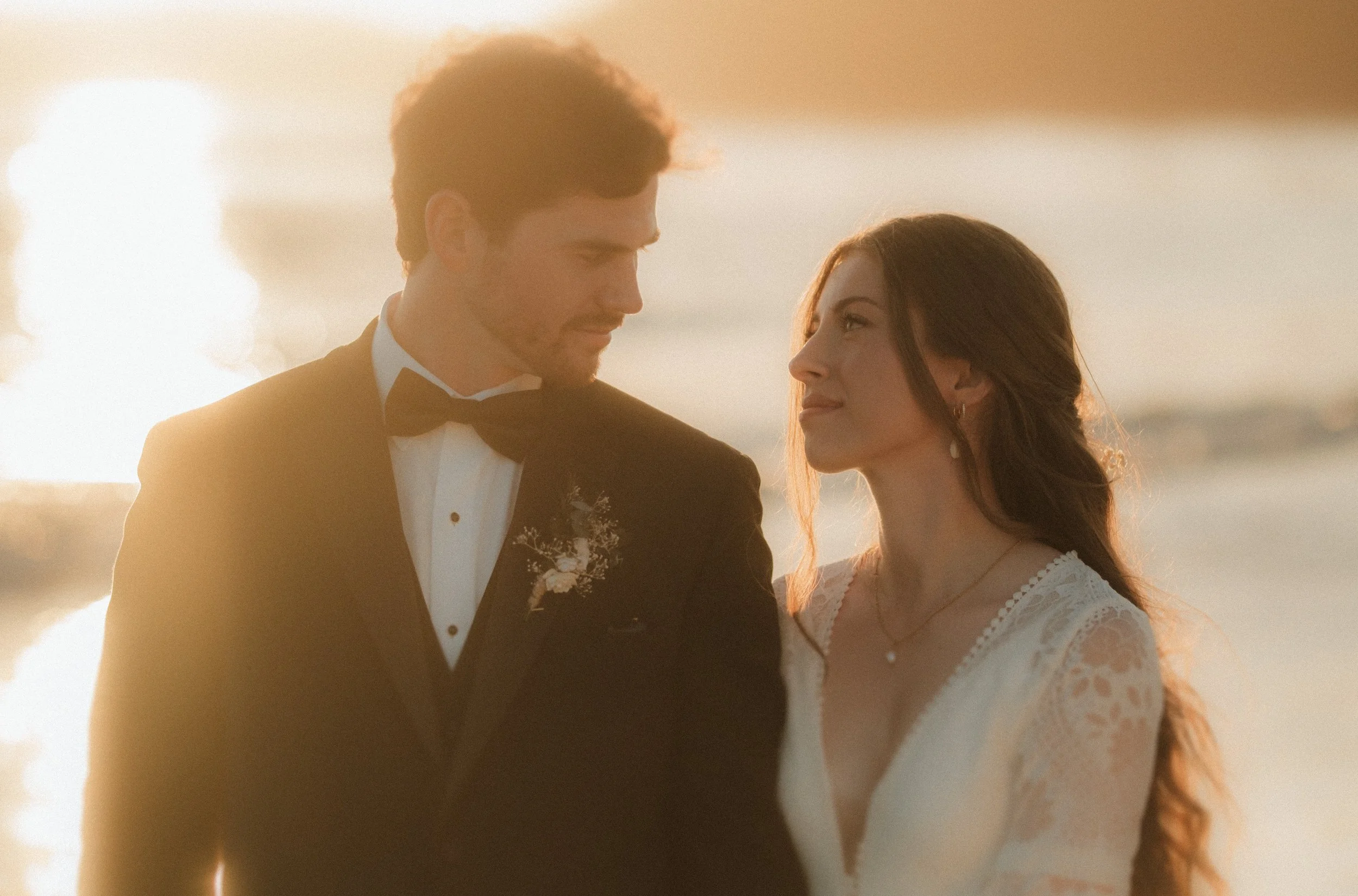 A man in a tuxedo and a woman in a white dress stand close together near a body of water during sunset, looking into each other's eyes in Tofino, BC