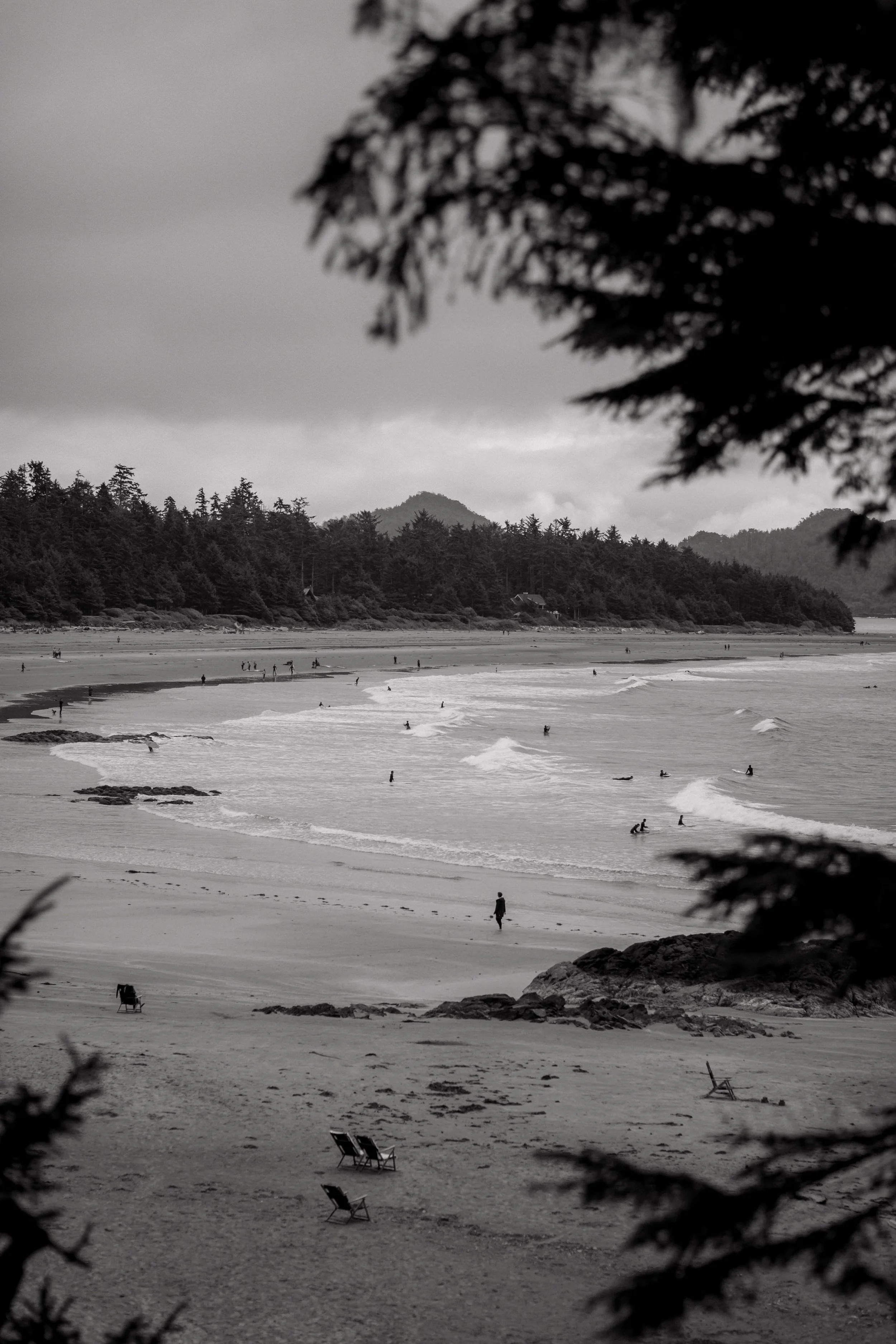 A black and white photo of a beach with people surfing and walking, viewed through trees from the Wickaninnish Inn, Tofino. 