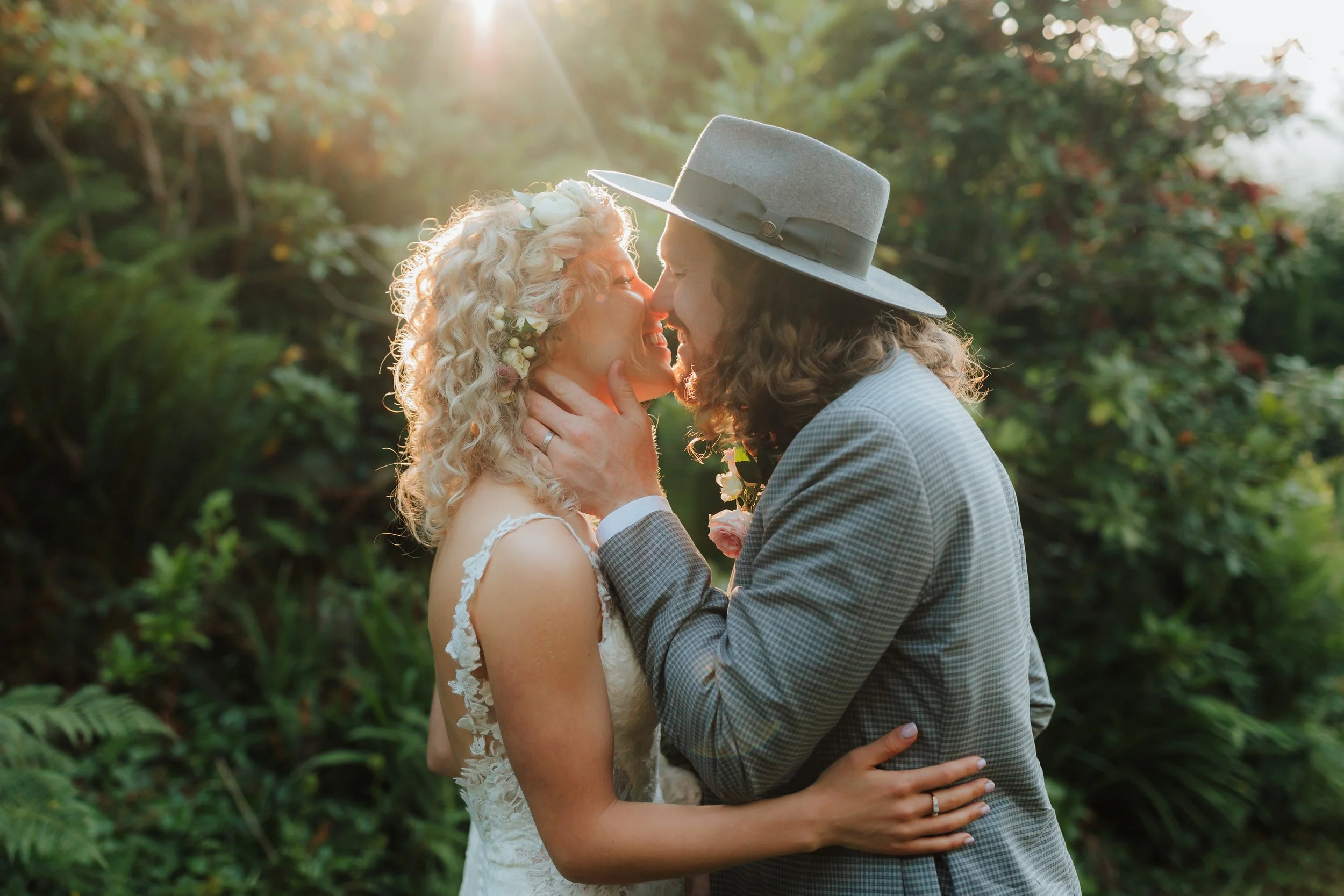 A couple in wedding attire sharing an intimate moment outdoors at sunset. The woman has curly blonde hair with flowers, and the man has long hair with a gray hat, holding her face gently.