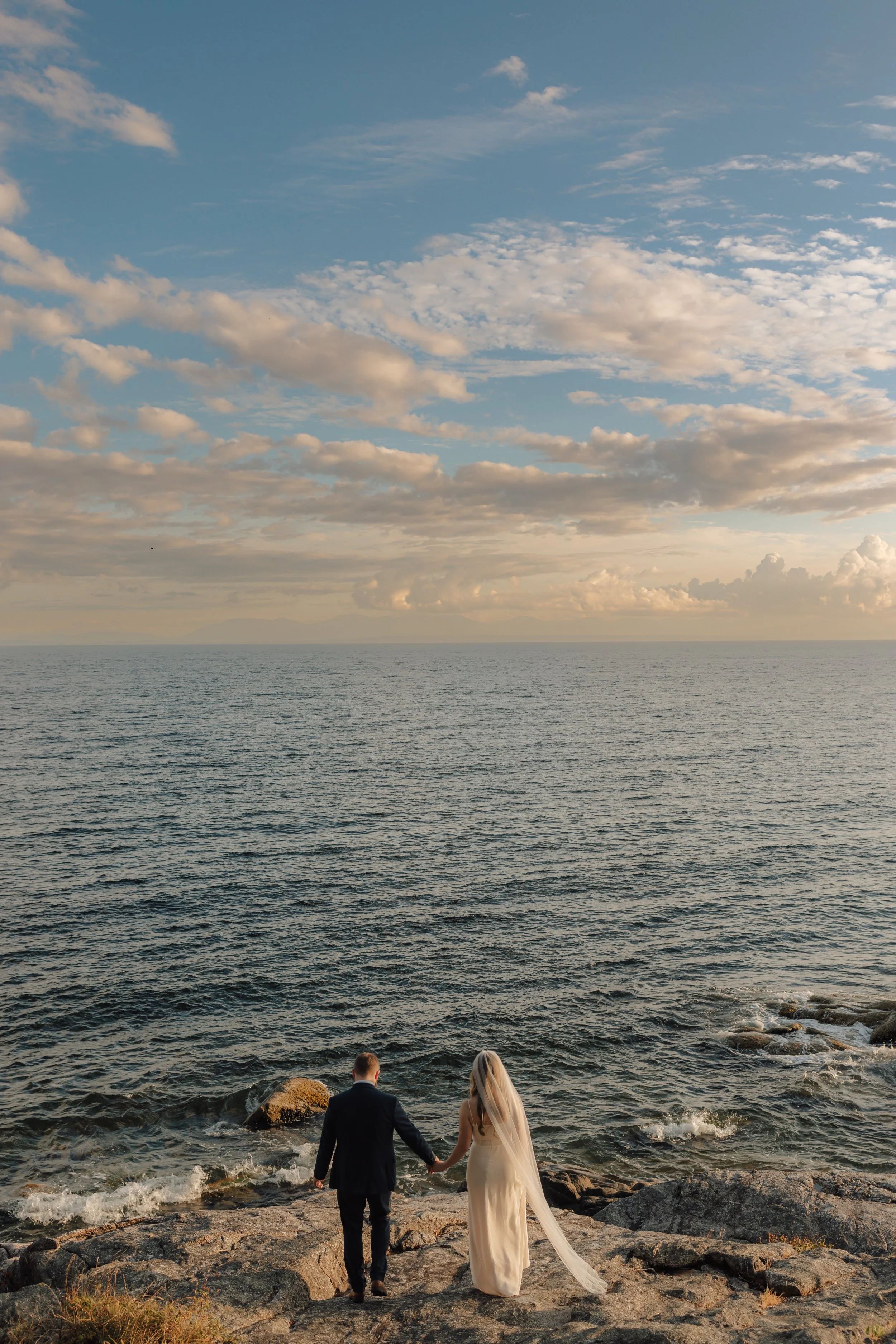 Elopement couple dressed in wedding attire holding hands and walking towards the ocean on a rocky shoreline during sunset, with clouds in the sky on the Sunshine Coast, BC