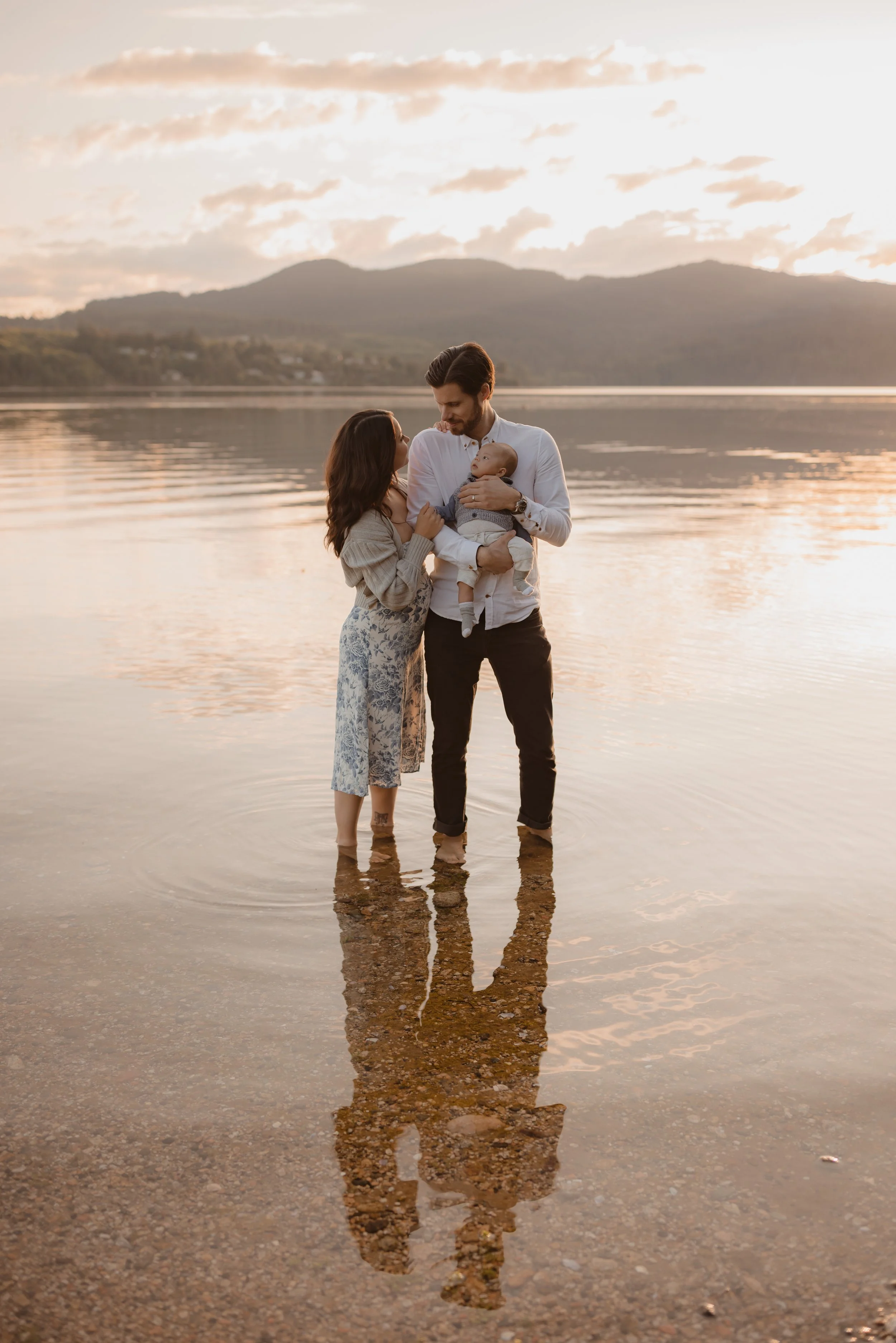 A family of three standing in shallow water at the edge of the ocean during sunset, with mountains in the background. The parents are holding their baby, and the reflection of the family is visible in the water on the Sunshine Coast.