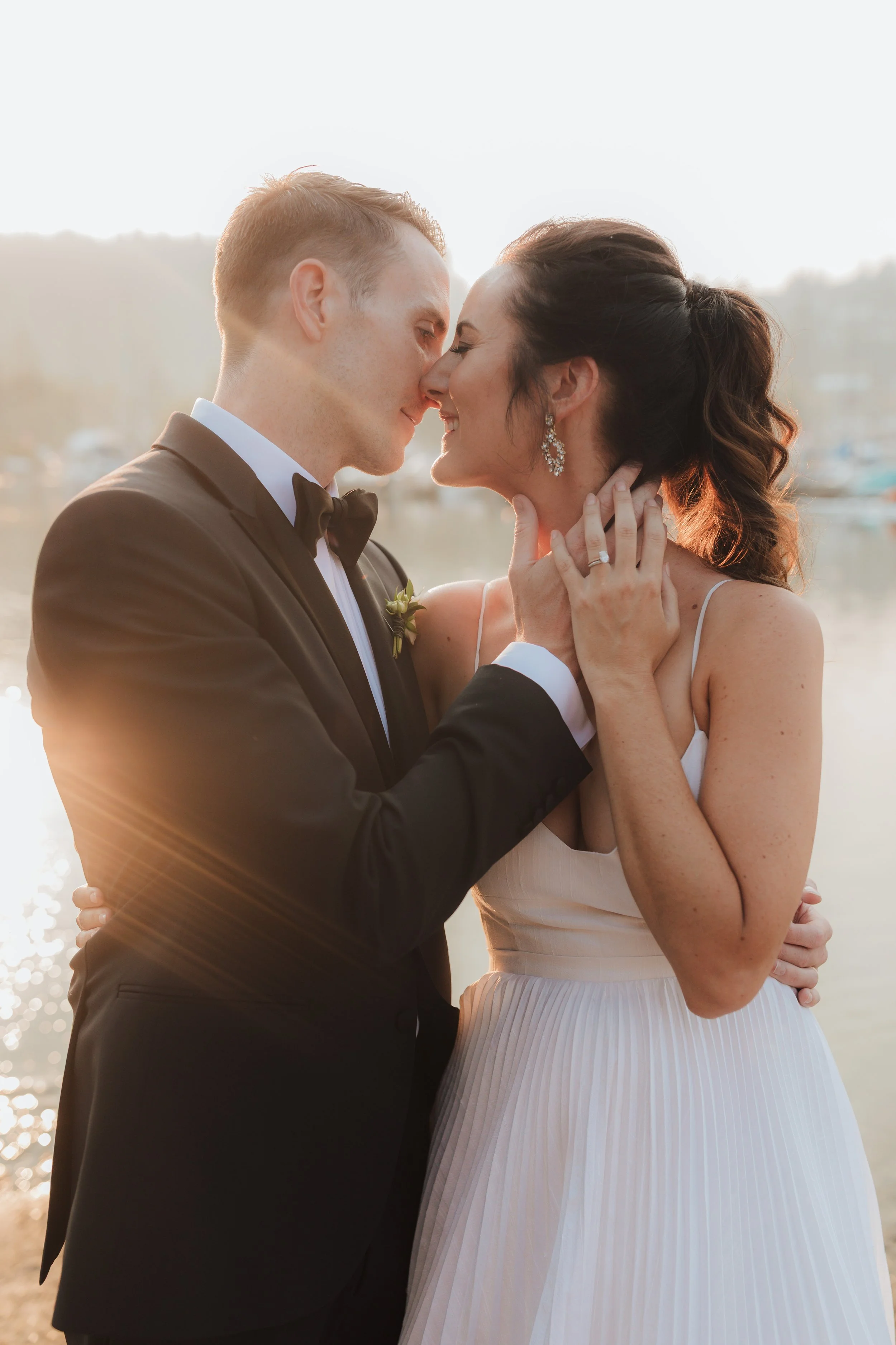 A newlywed couple shares a close, intimate moment by a waterfront, with the sunset casting a warm glow around them.