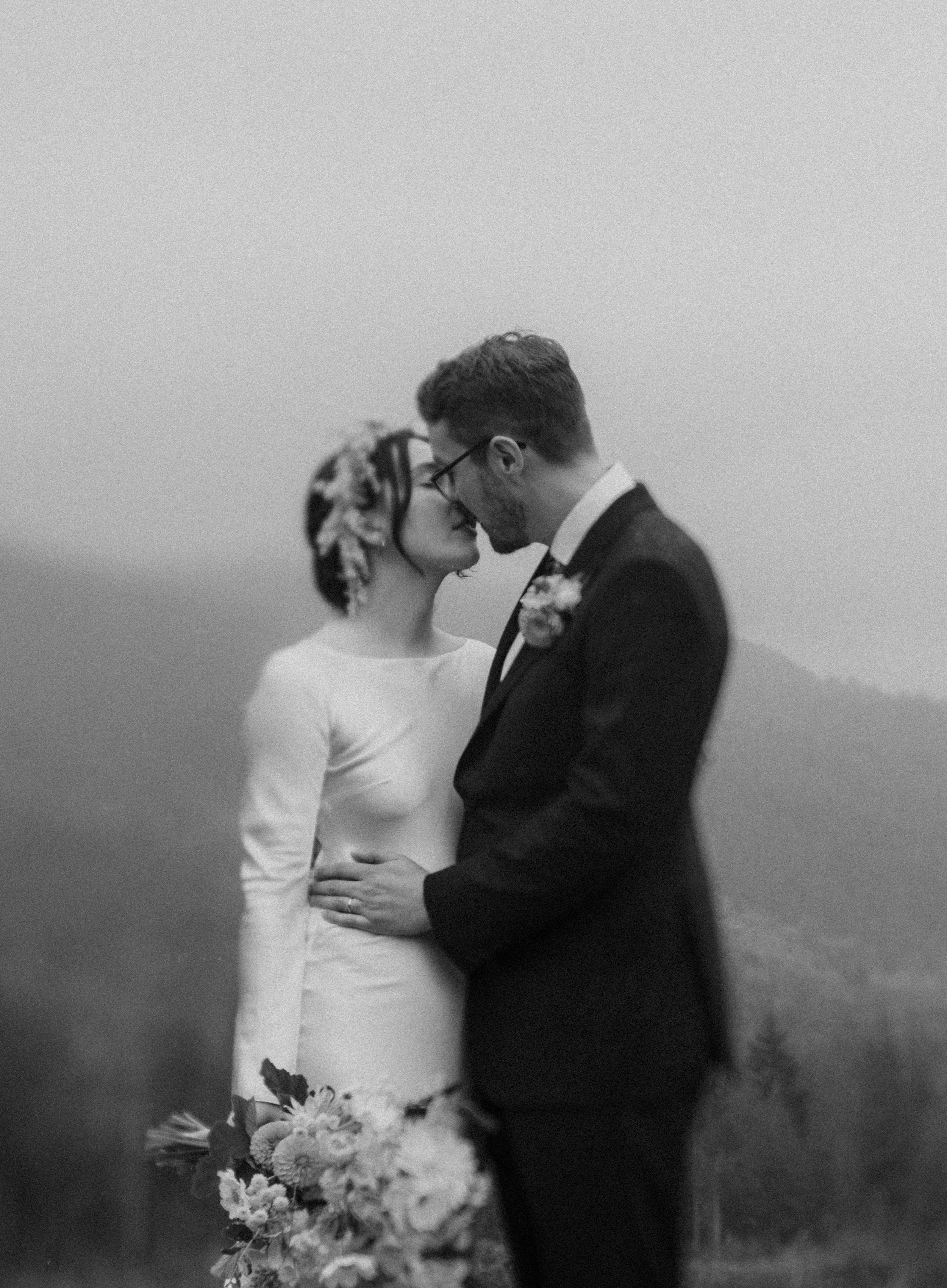 Black and white photo of a bride and groom standing close together, noses touching, outdoors with a foggy or misty background.