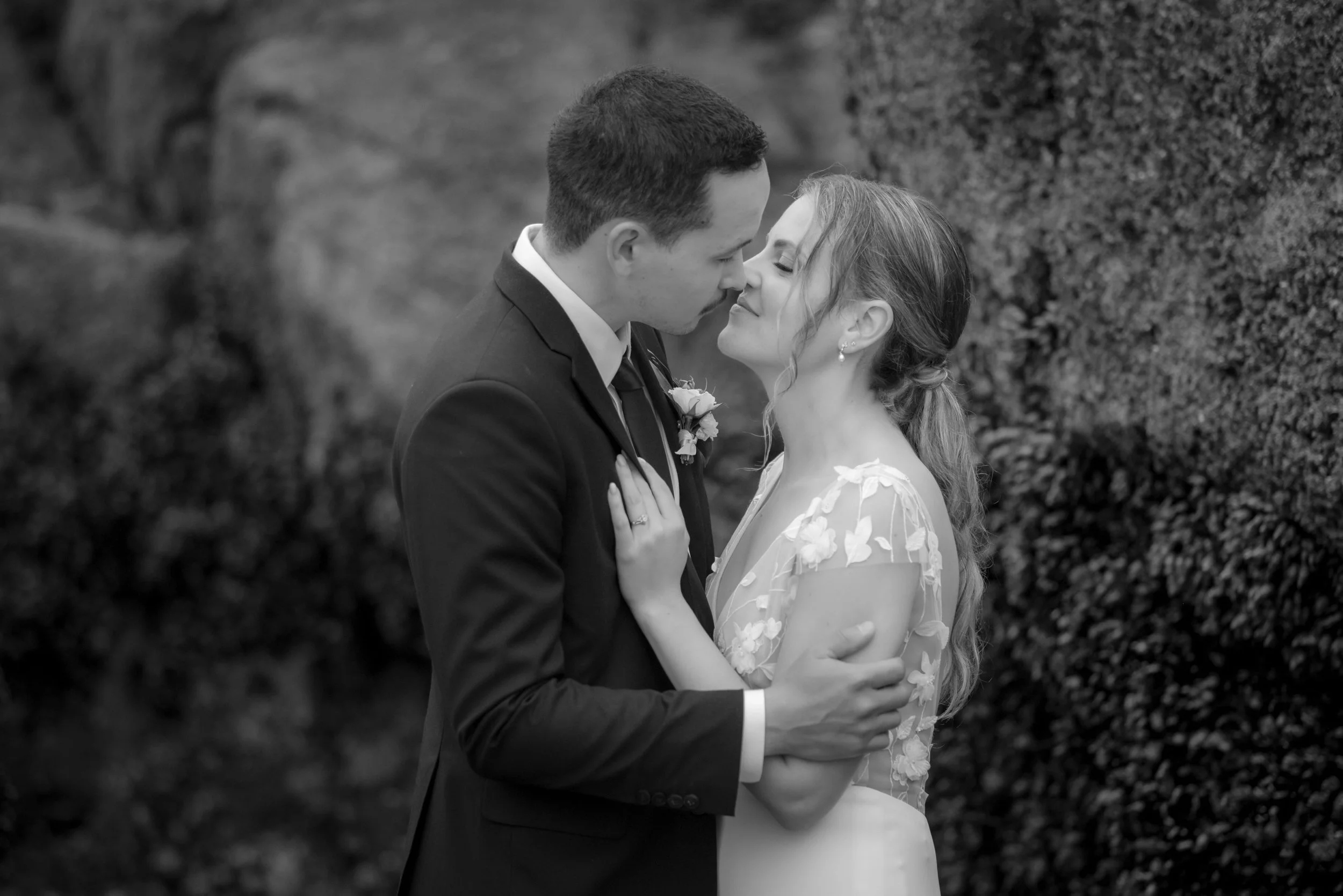 Black and white photo of a wedding couple close together, about to kiss, outdoors with bushes in the background.