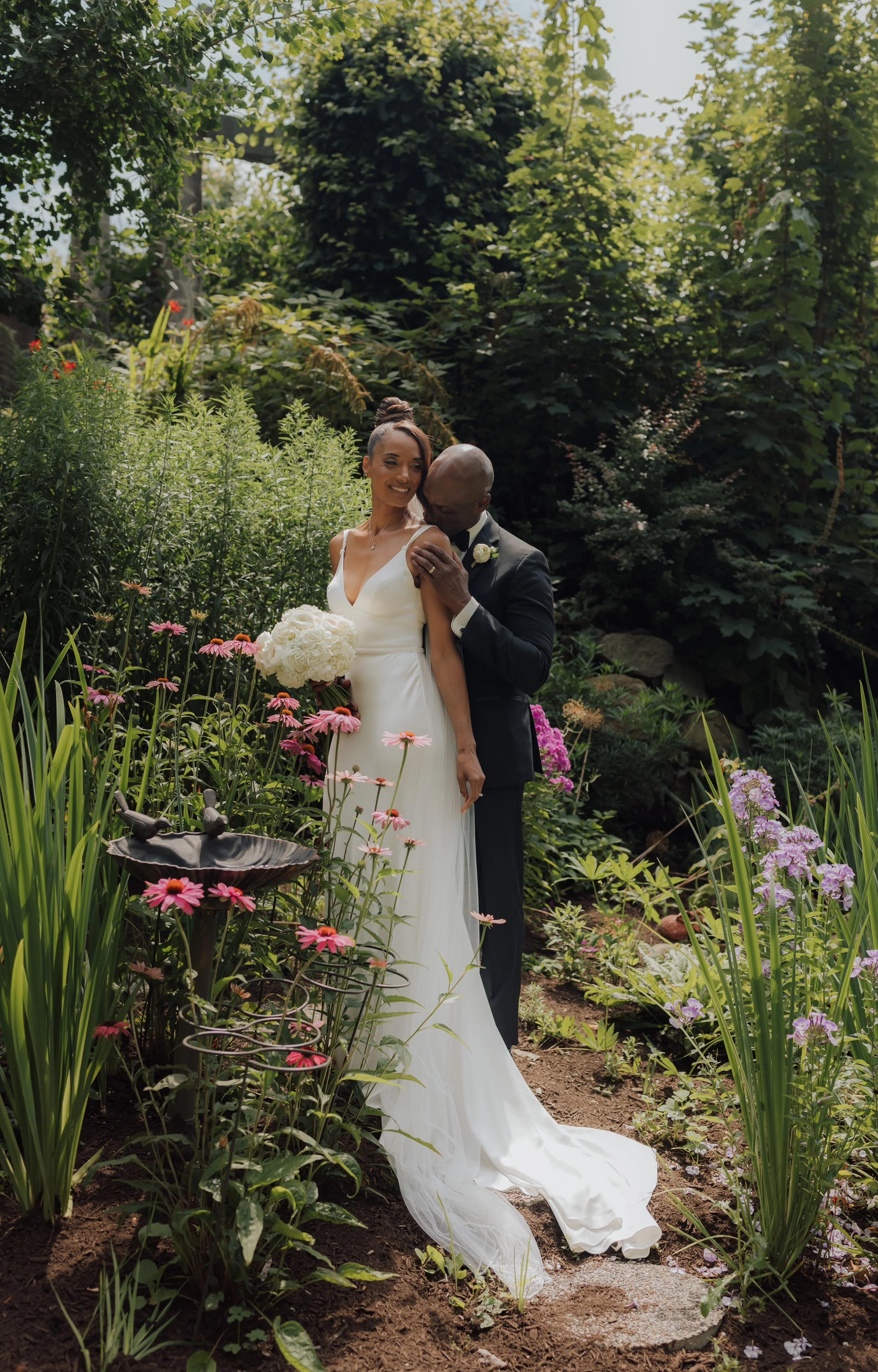 A newlywed couple stands in a lush garden surrounded by pink and purple flowers, with the man embracing the woman and she holding a bouquet of white flowers at Hycroft Manor in Vancouver