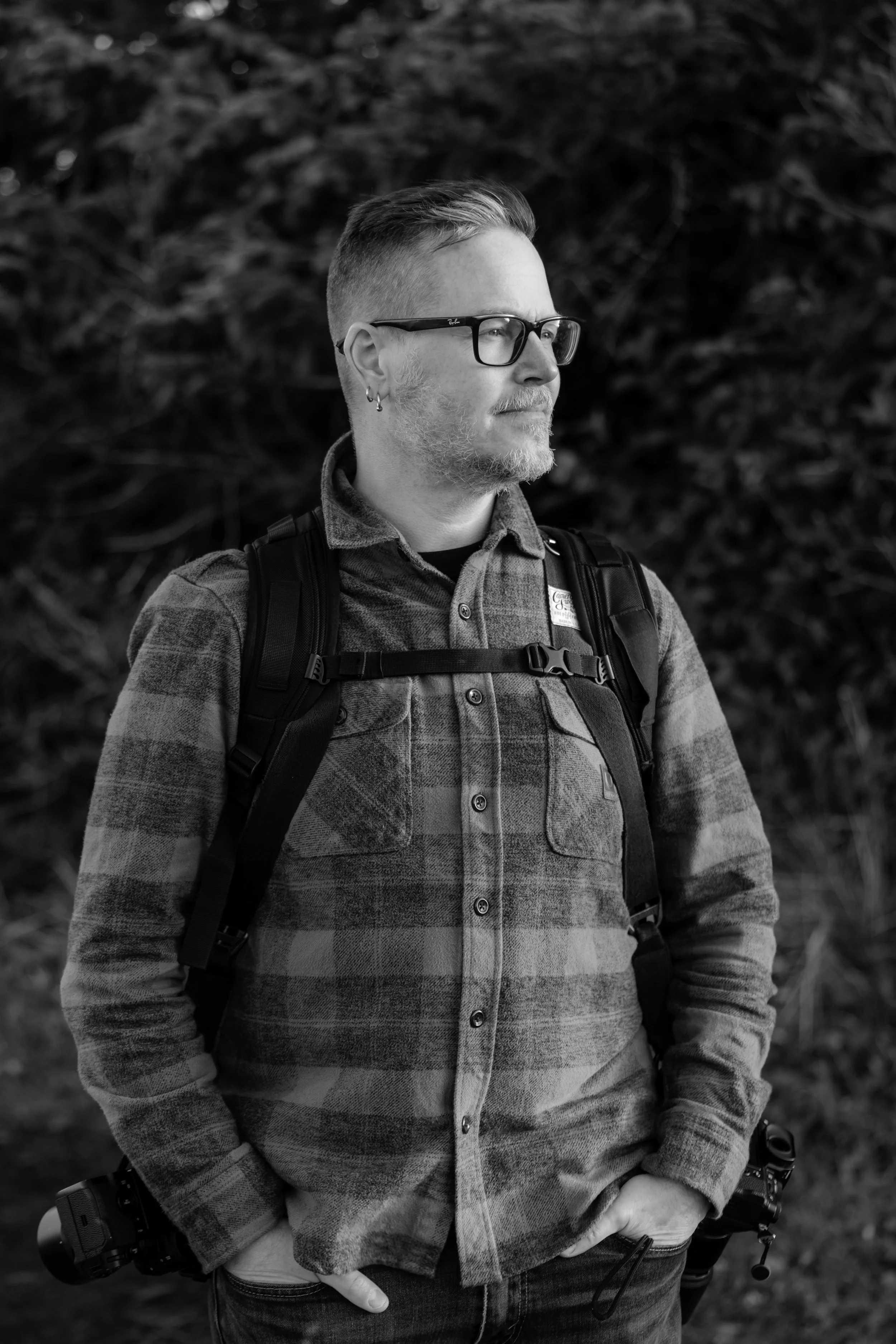 A man with glasses, beard, and earrings, wearing a plaid jacket and backpack, standing outdoors with a serious expression.