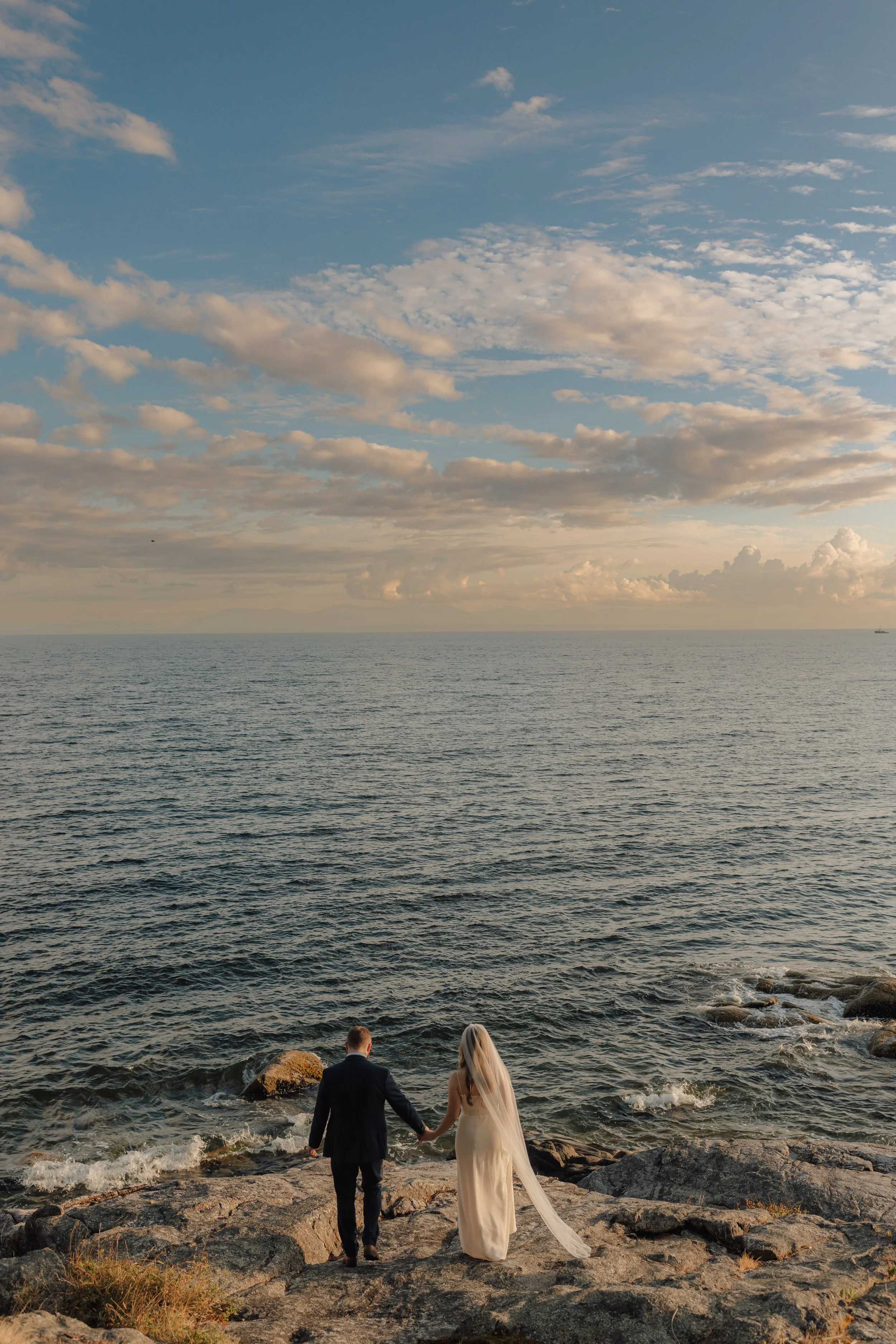 A bride and groom walking hand in hand along a rocky shoreline at sunset, with the ocean and a partly cloudy sky in the background at golden hour on the Sunshine Coast.