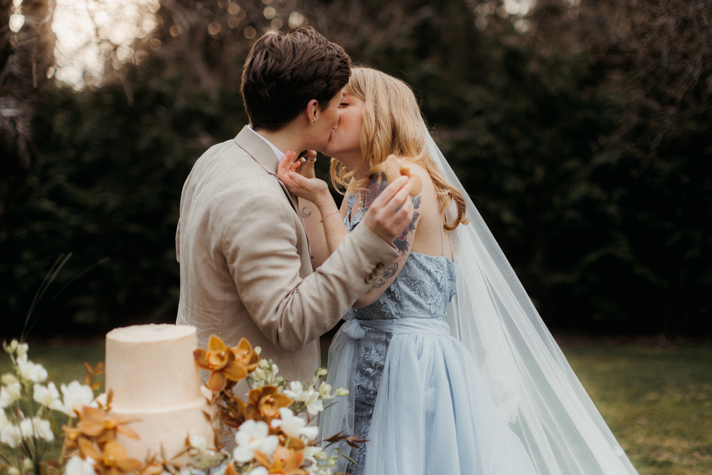 A couple sharing a kiss at their outdoor wedding, with a wedding cake and floral arrangement in the foreground on the Sunshine Coast.
