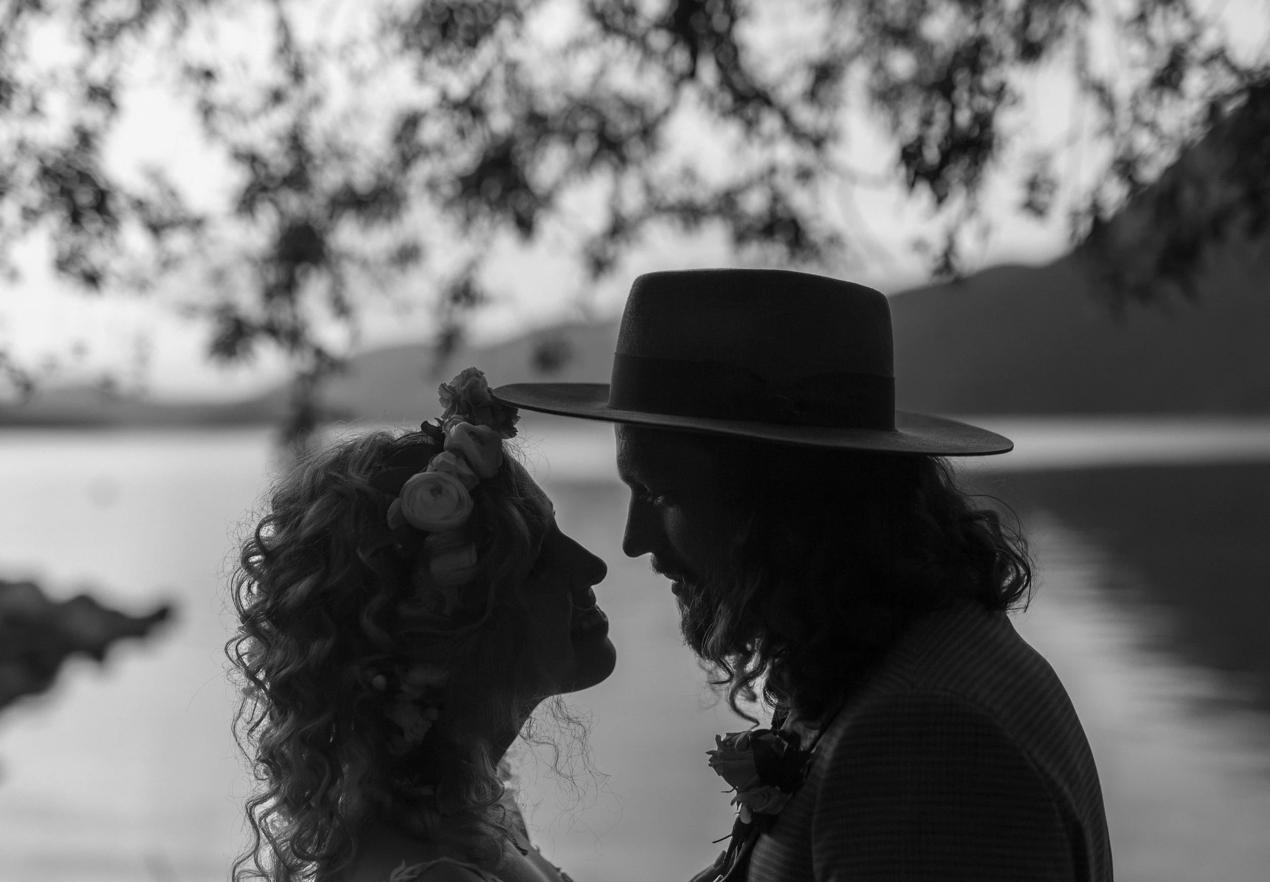 Silhouette of a couple facing each other near a body of water, with trees and hills in the background, during sunset or sunrise.