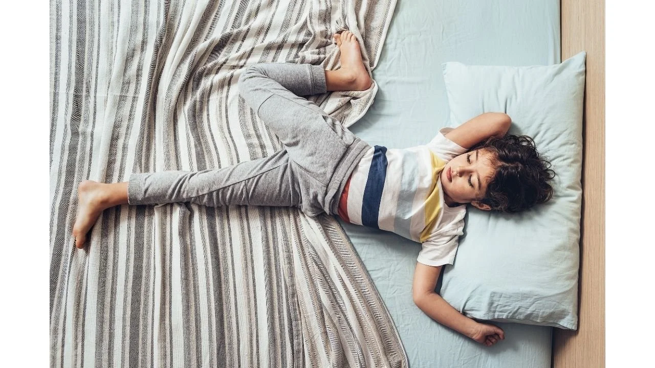 A young child with curly hair sleeping on a bed with a light blue pillow and striped bedsheet, lying on their back with arms behind their head.