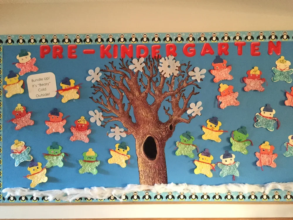 Pre-kindergarten classroom bulletin board with a large paper tree in the center with orca whale border at the top and bottom, and cotton batting at the bottom representing snow. Colorful felt snowmen wearing hats and scarves are attached around the t