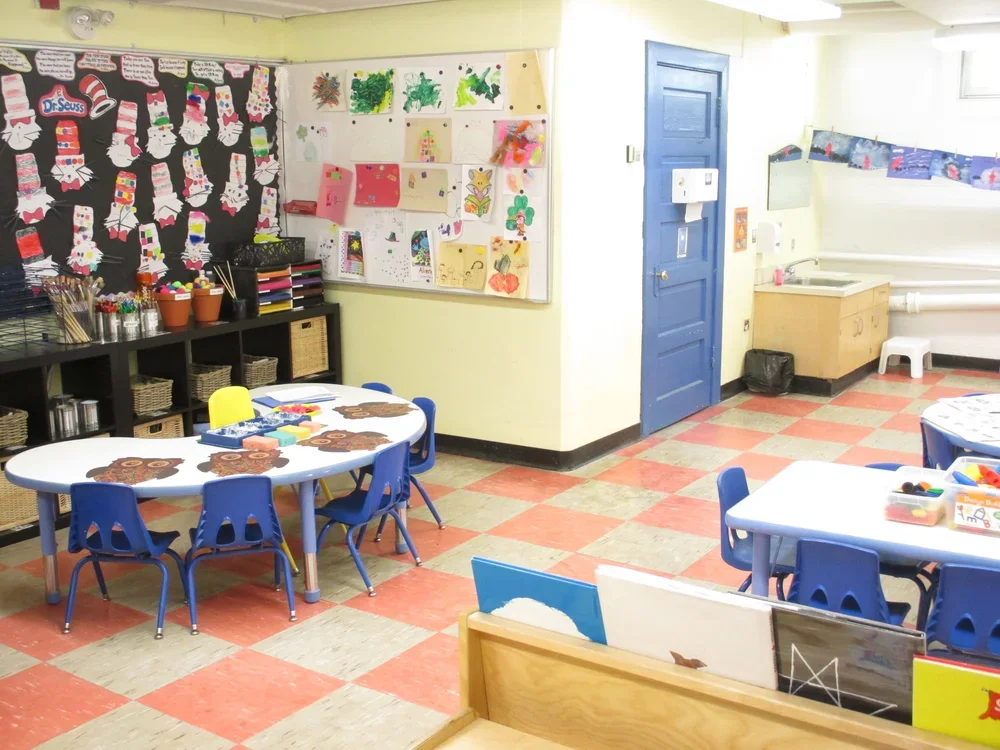 A classroom with small tables and chairs, art projects on the wall, art supplies on shelves, and colored tiled flooring.