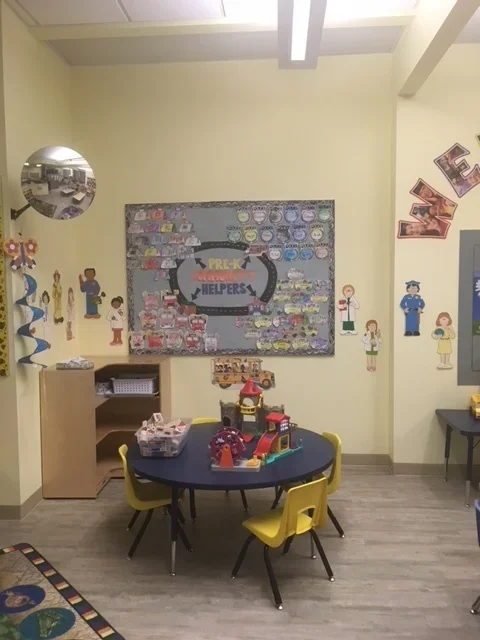 A classroom with a round table, yellow chairs, and shelves, decorated with posters and artwork related to preschool helpers and the letter V.