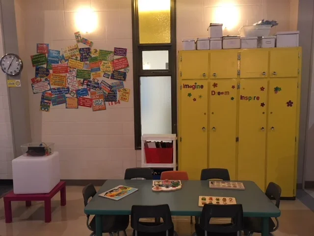 A classroom corner with yellow lockers decorated with the words "Imagine," "Dream," and "Inspire" and colorful flower stickers, a table with chairs and reading materials, a white storage box on a small pink table, a bulletin board with motivational q