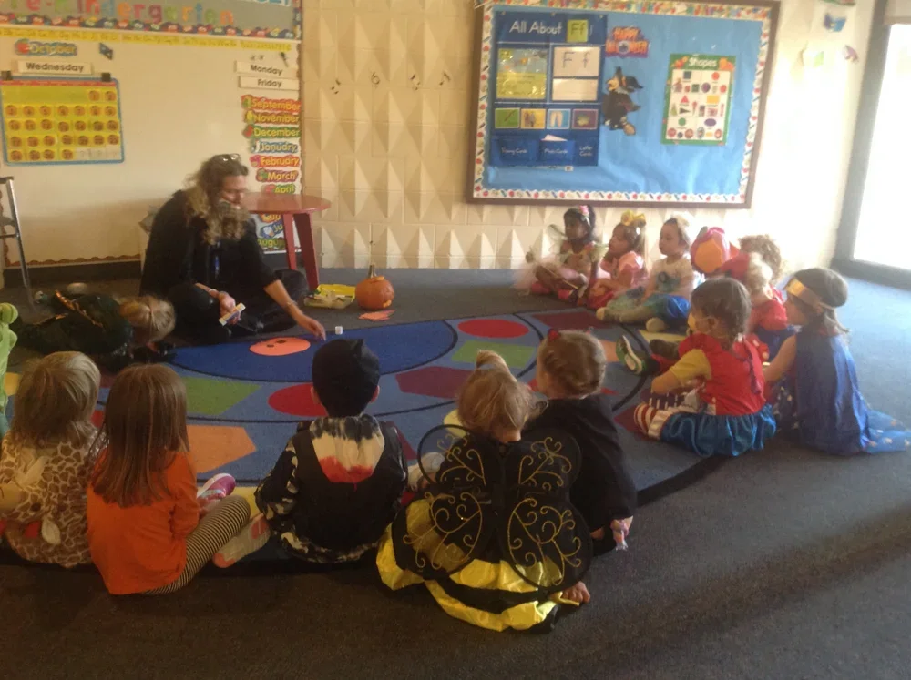 Kindergarten classroom with children sitting on a colorful circular rug, listening to a teacher who is seated on the floor with a pumpkin and teaching materials, colorful posters and calendar on the wall.