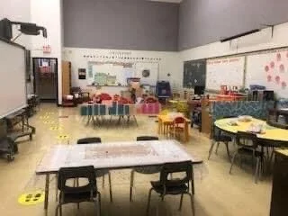 Empty classroom with tables and chairs, colorful decorations, and educational materials on the walls and chalkboard.