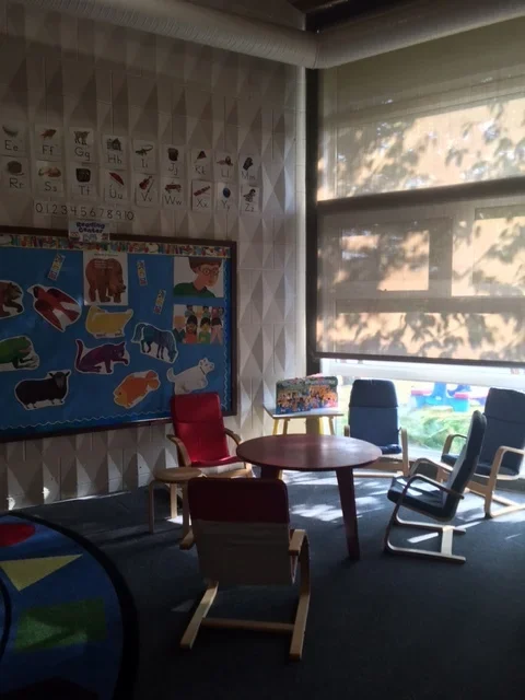 Classroom corner with chairs around a small round table, a window with shade partially down, and educational posters on the wall, including animal pictures and alphabet charts.