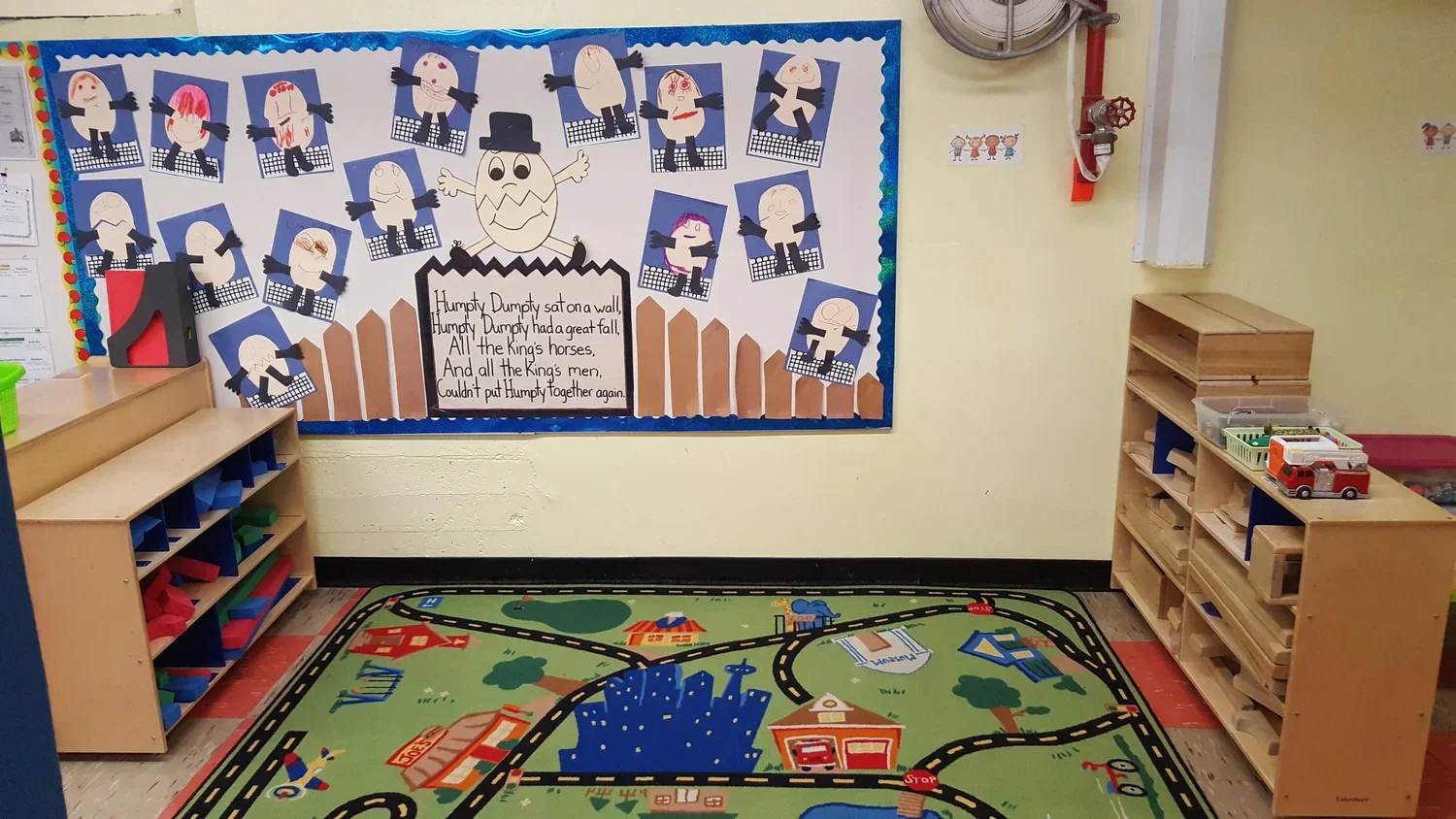 Classroom corner with a colorful play rug depicting roads and buildings, storage shelves with toys, and a bulletin board with Halloween-themed artwork featuring paper crafts of Frankenstein's monster heads and a poster of Humpty Dumpty.