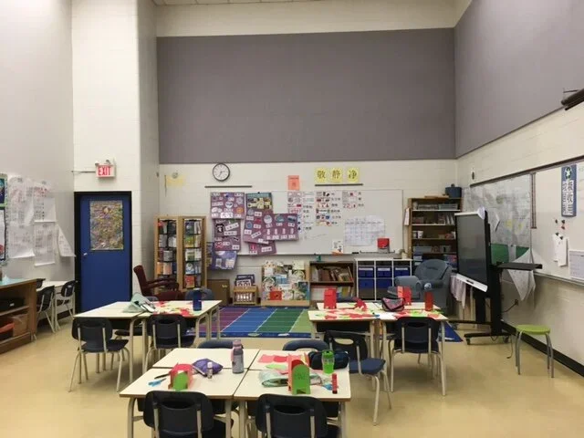 Empty classroom with desks set for a meal, decorated with colorful paper houses, in a school or community center.