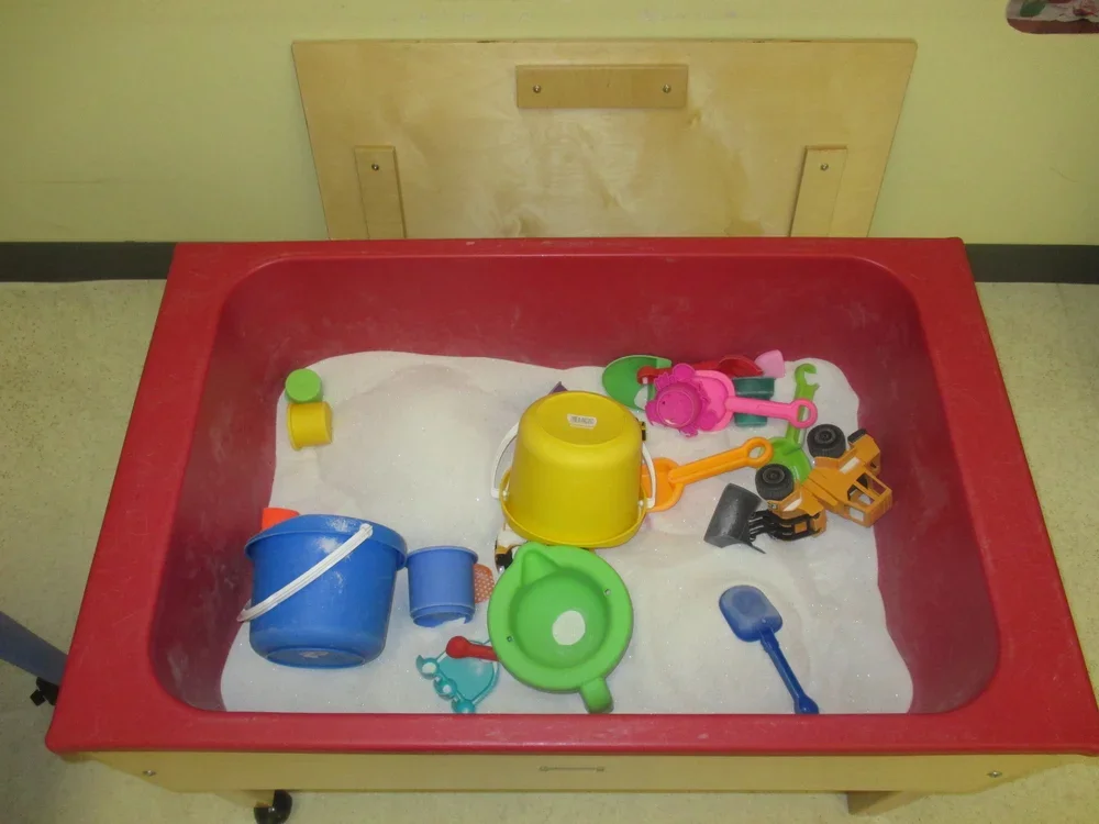 Toy sandbox with plastic buckets, shovels, and other sand toys, filled with white sand.